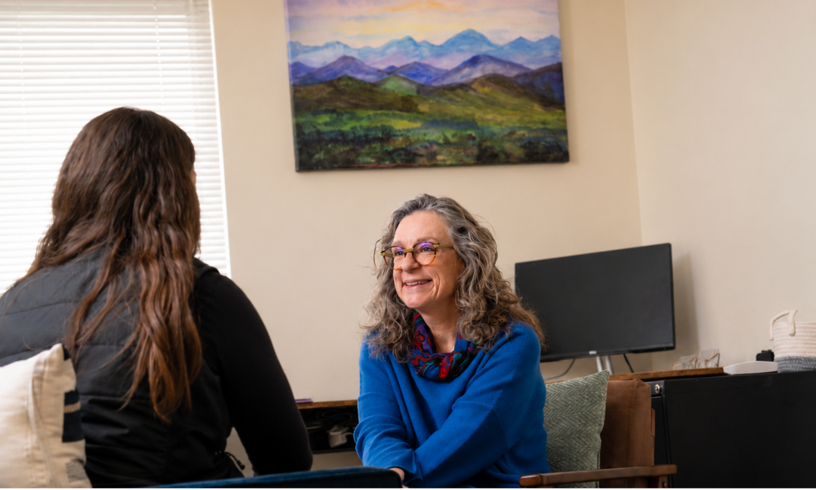 A woman with glasses and gray curly hair smiling and talking to another woman with long brown hair in an office setting.