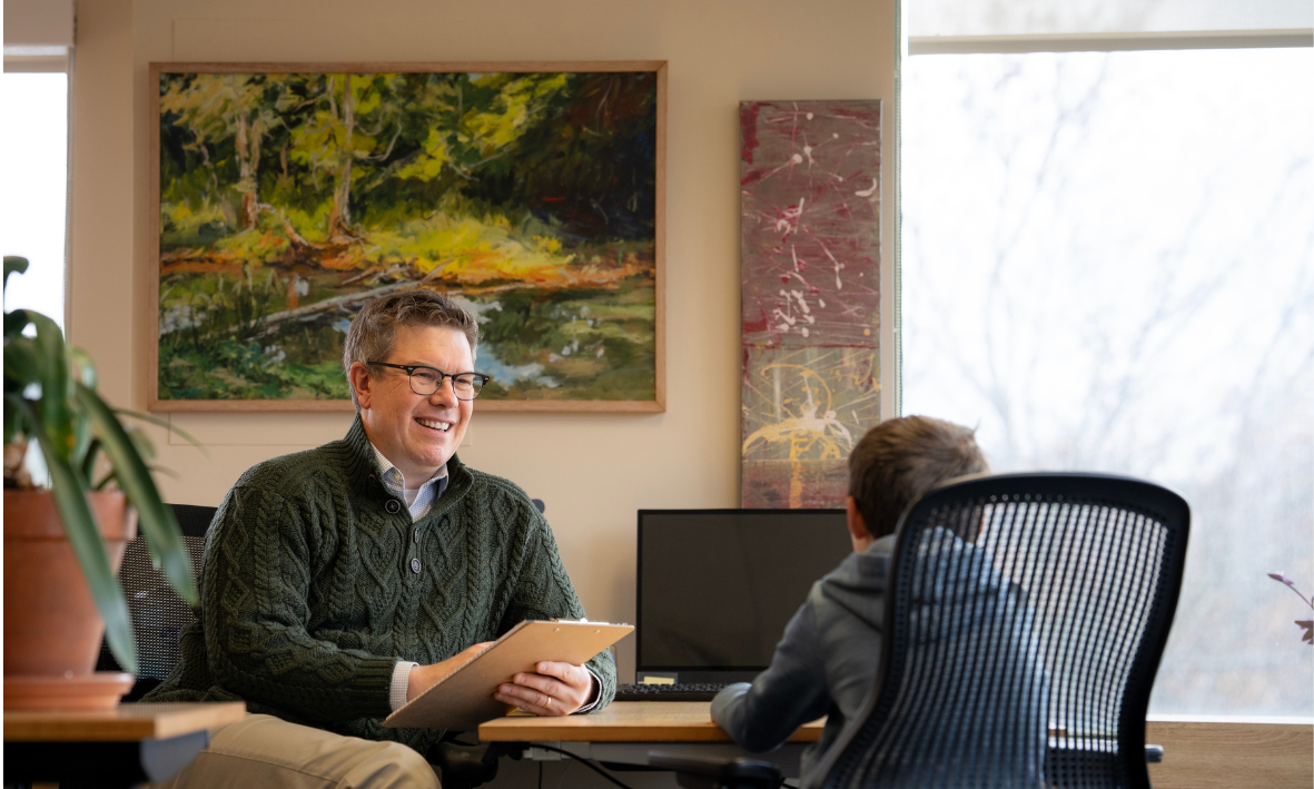 A man with glasses and a green sweater smiling while talking to a student in an office with a window, paintings on the wall, and a plant on a desk.