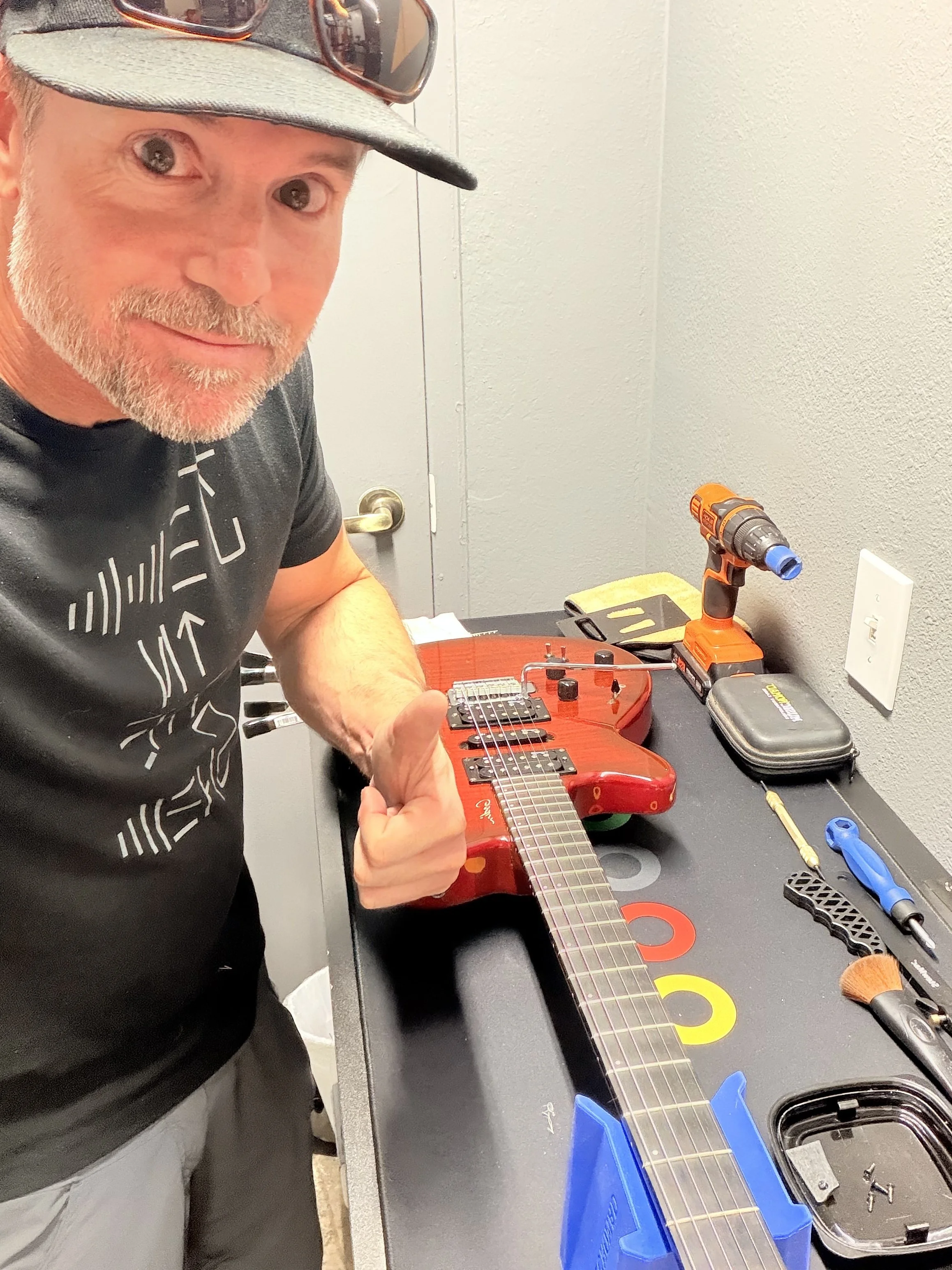 A man with a beard wearing a baseball cap and sunglasses on top, working on an electric guitar in a repair workspace. The workspace has various tools, a cordless drill, brushes, and other parts on a black workbench.