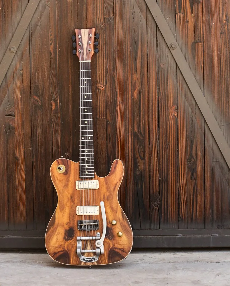 A vintage-style electric guitar with a natural wood finish leaning against a wooden barn door.
