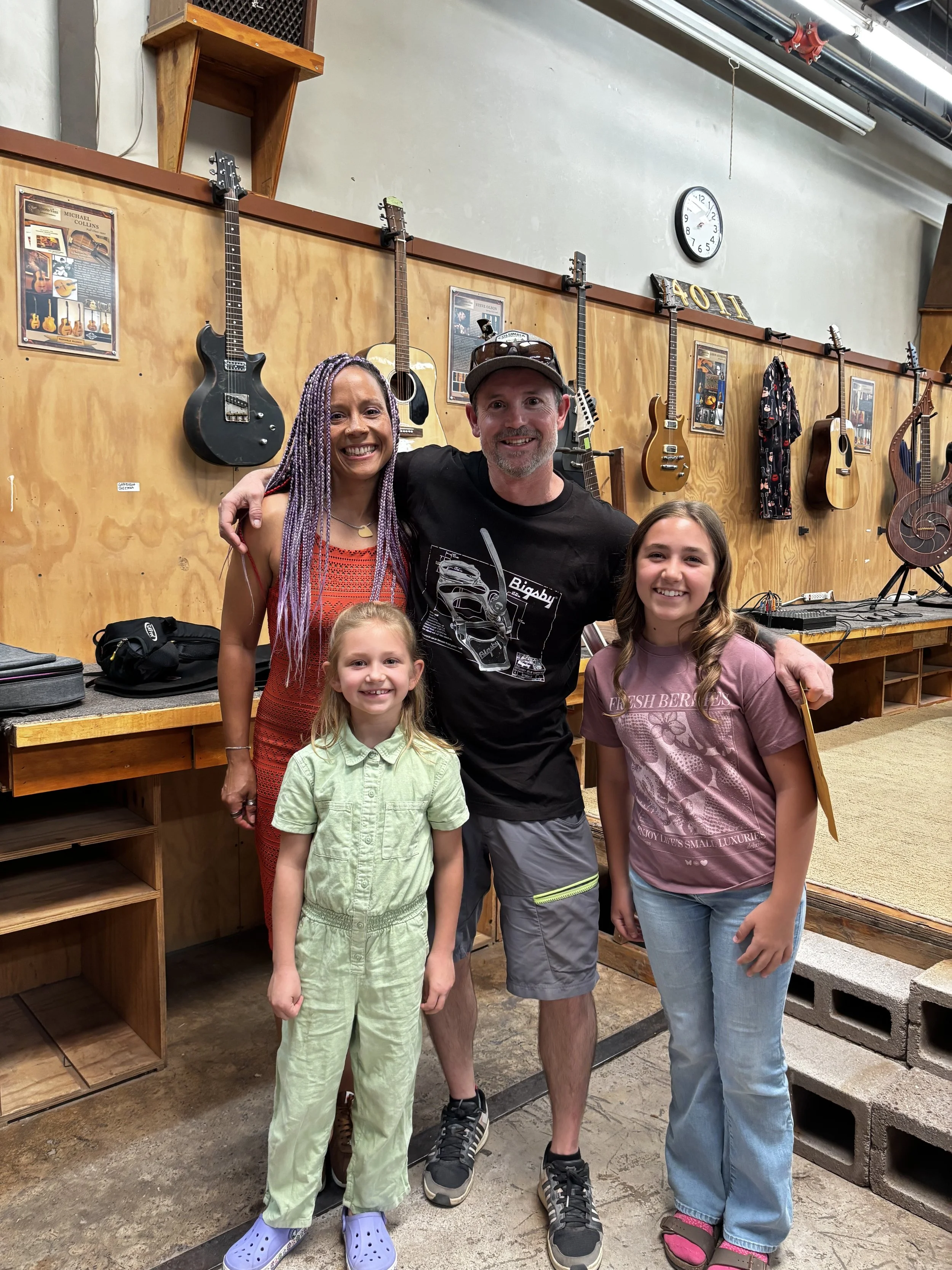 A group of five people, including two children and three adults, smiling and posing together indoors with musical instruments hanging on the wall behind them.
