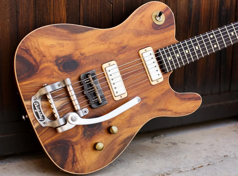 Electric guitar with a natural wood grain finish, black fretboard, and cream pickups, resting against a wooden wall.