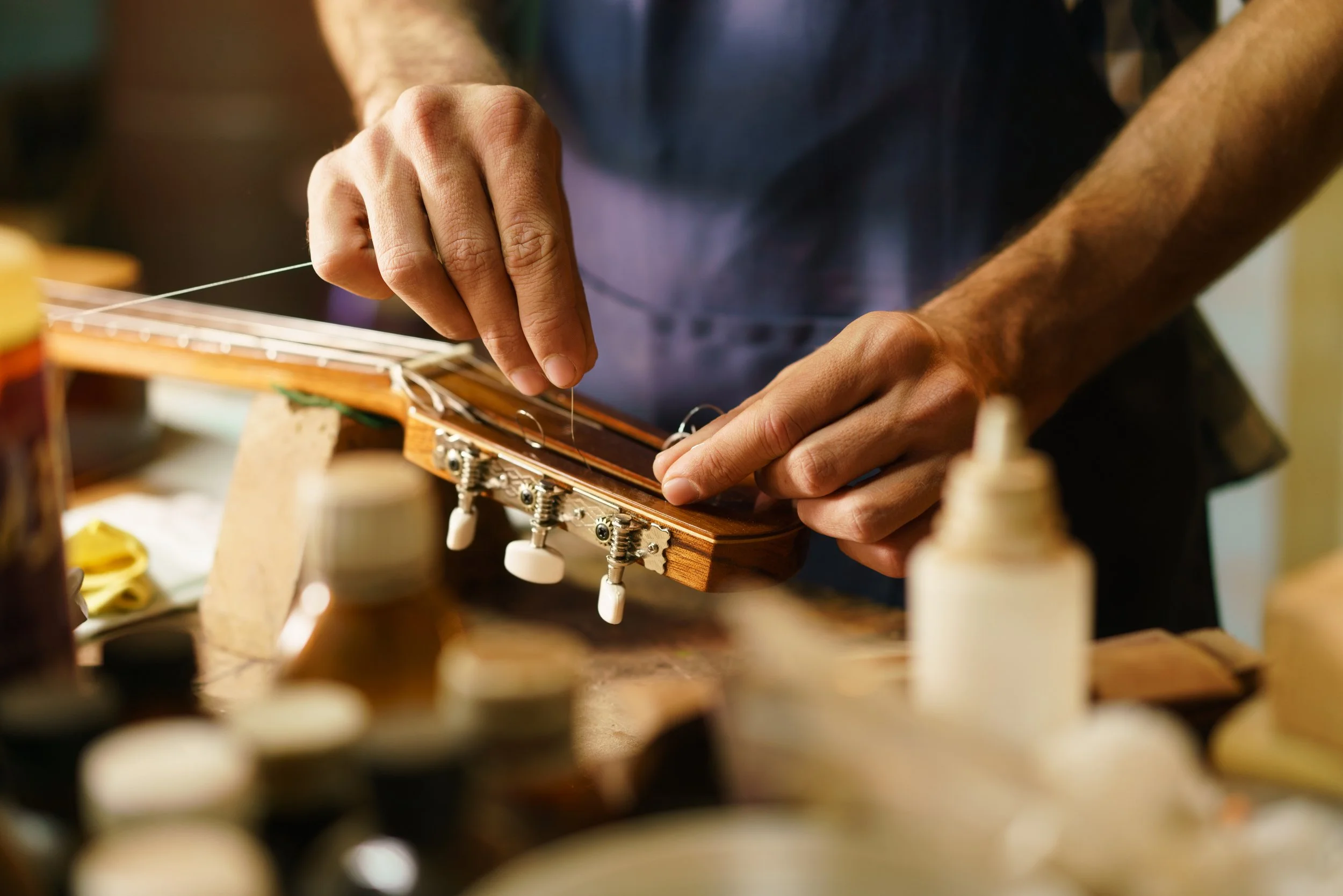 Close-up of a person's hands threading a string through the tuning pegs of a guitar neck, with various objects blurred in the foreground.