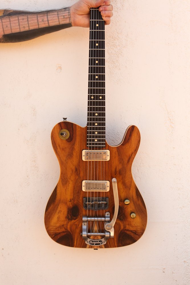 A person holding a vintage-style electric bass guitar with a wooden body against a white wall.