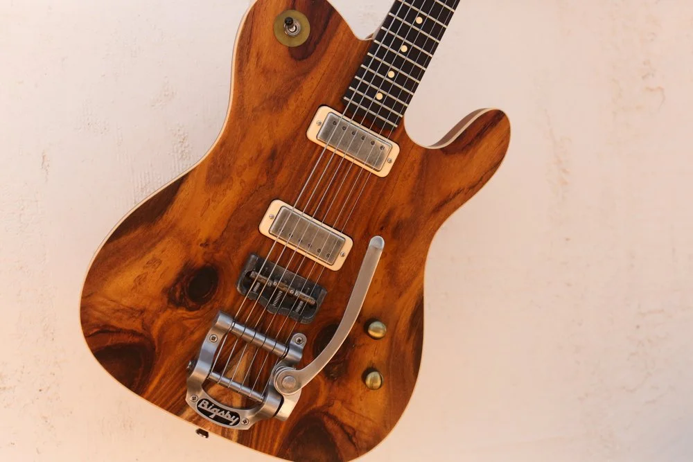 A wooden electric guitar with a natural finish, featuring two humbucker pickups, a Bigsby tremolo, and black fingerboard with dot inlays, hanging against a light-colored wall.