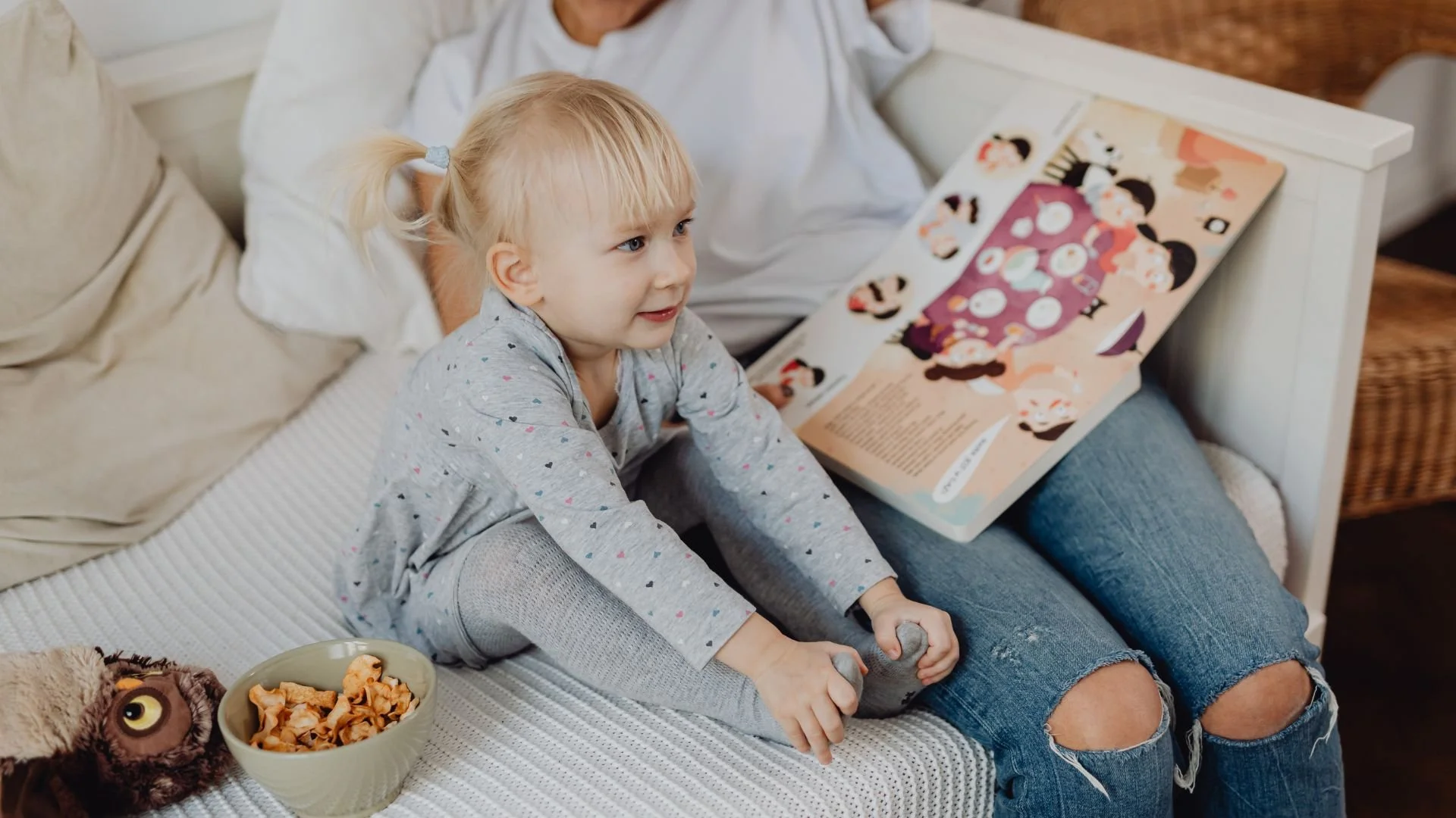 A young girl sitting on a bed, looking at a book, with a bowl of snacks and a stuffed owl nearby.