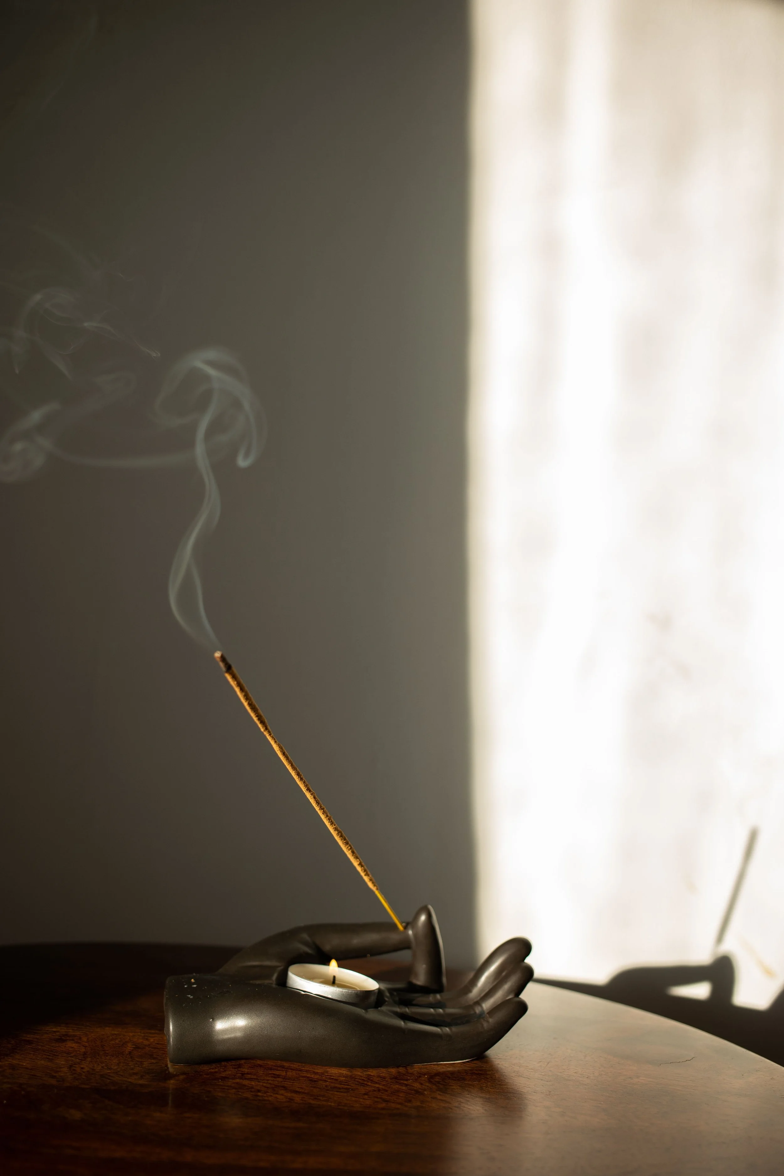 A lit tea light candle in a black holder and a person's gloved hand holding a long matchstick with a burning tip, with a shadowed background and a left-placed bright light source casting shadows.