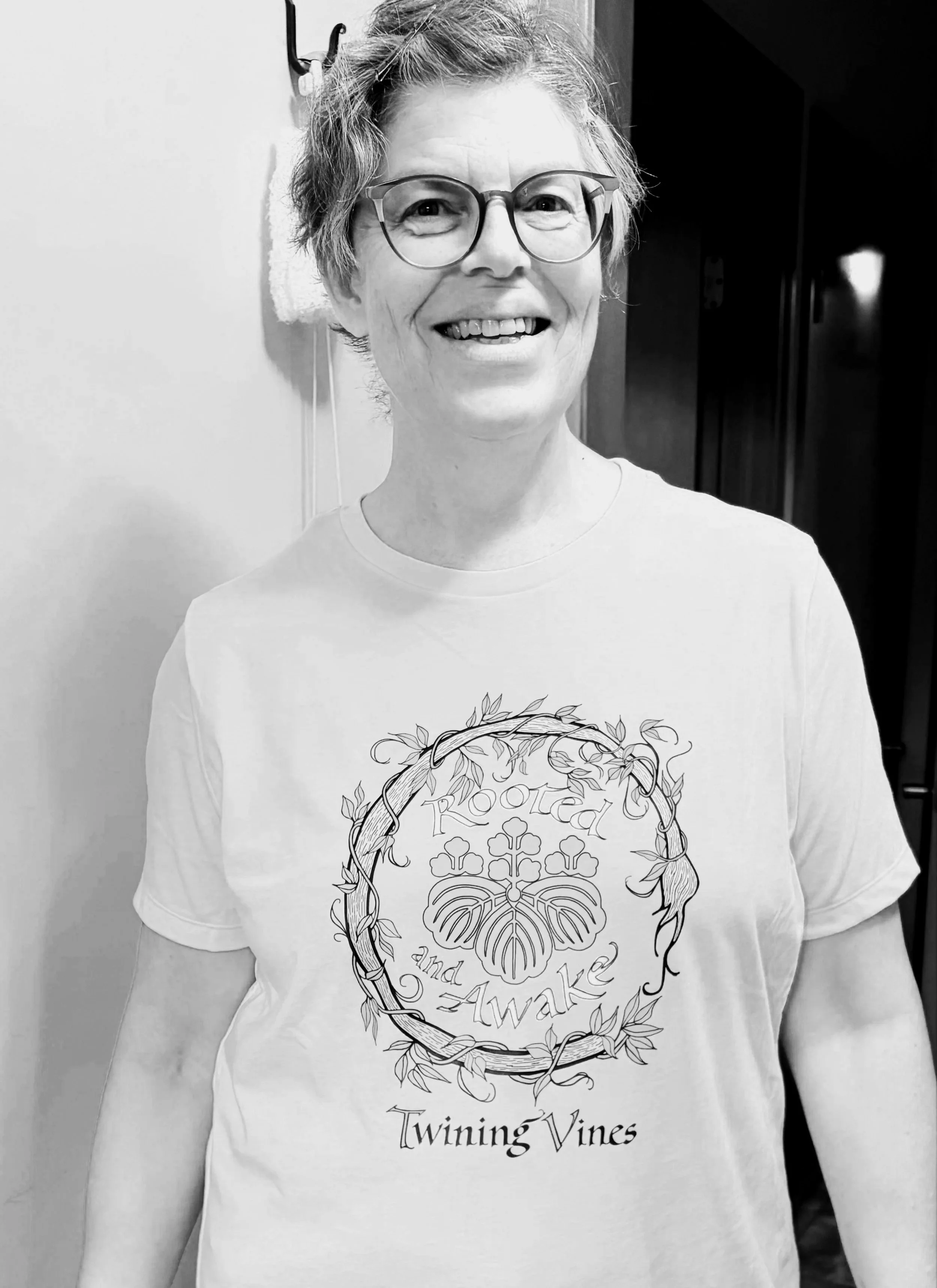 A smiling woman with short hair and glasses wearing a white T-shirt with a graphic design of vines and the text 'Twinning Vines'.