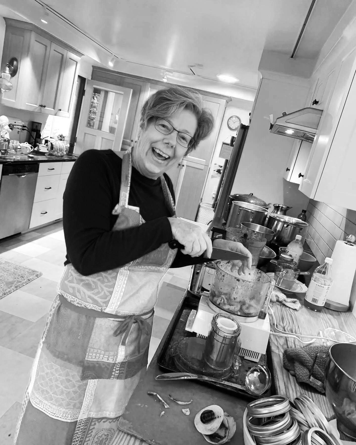 Joan Bateman, a smiling woman with glasses, wearing an apron, is cooking in a kitchen. She is chopping or blending something in a glass bowl placed on a table filled with various kitchen items and appliances.