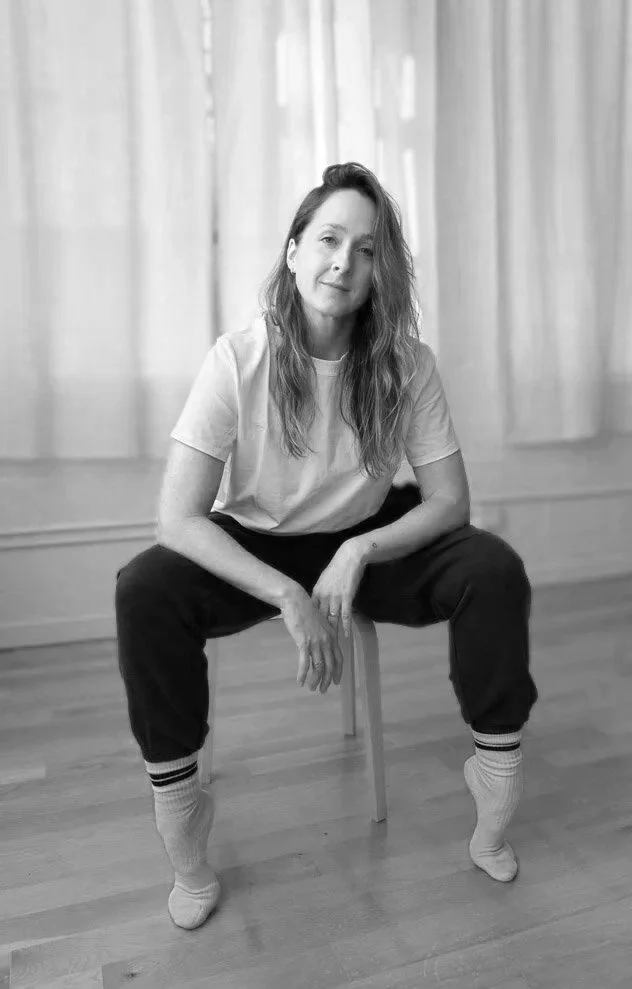 Court Morgan, a Walla Walla yoga teacher and young woman with wavy hair sitting on a small chair in a room with curtains, wearing a t-shirt, sweatpants, and socks, looking at the camera in a relaxed pose.