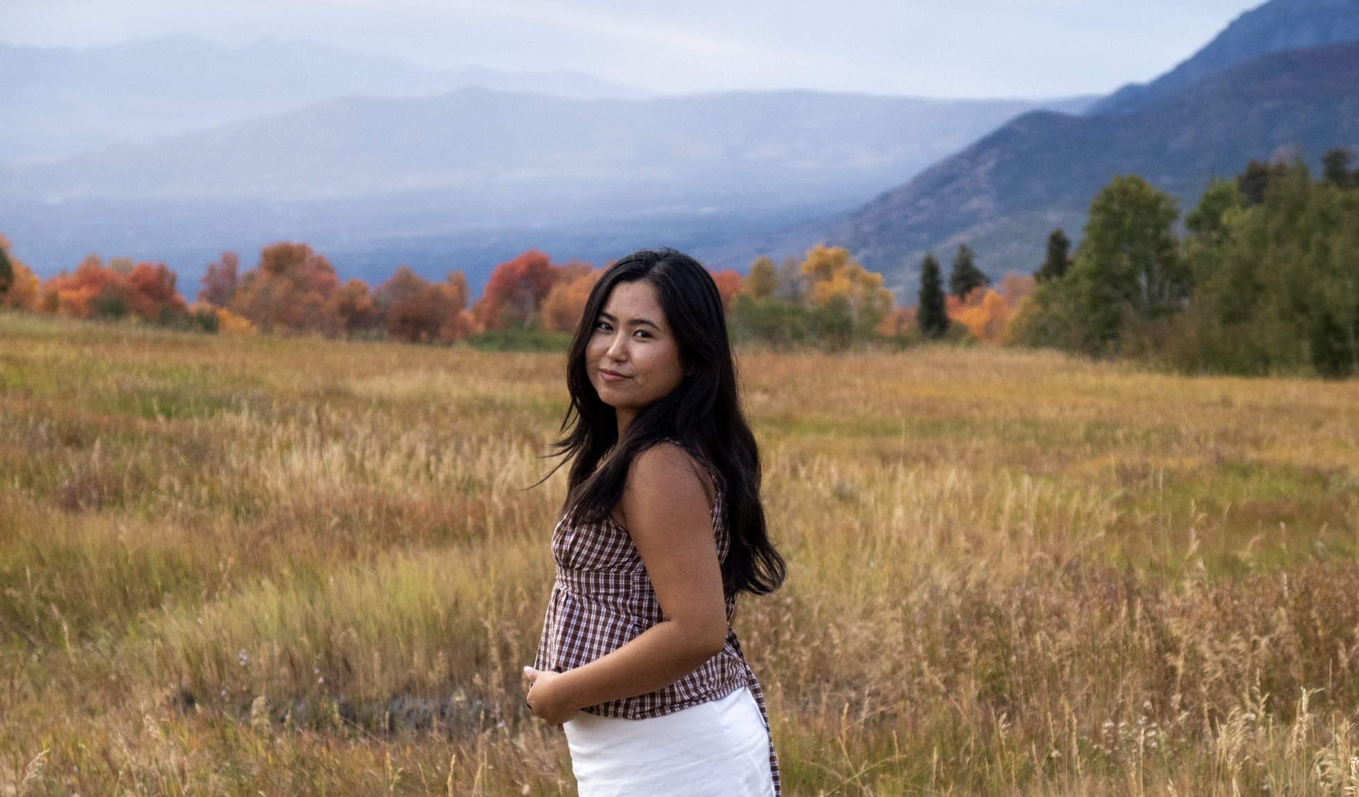 A woman standing in a grassy field with colorful autumn trees and mountains in the background.