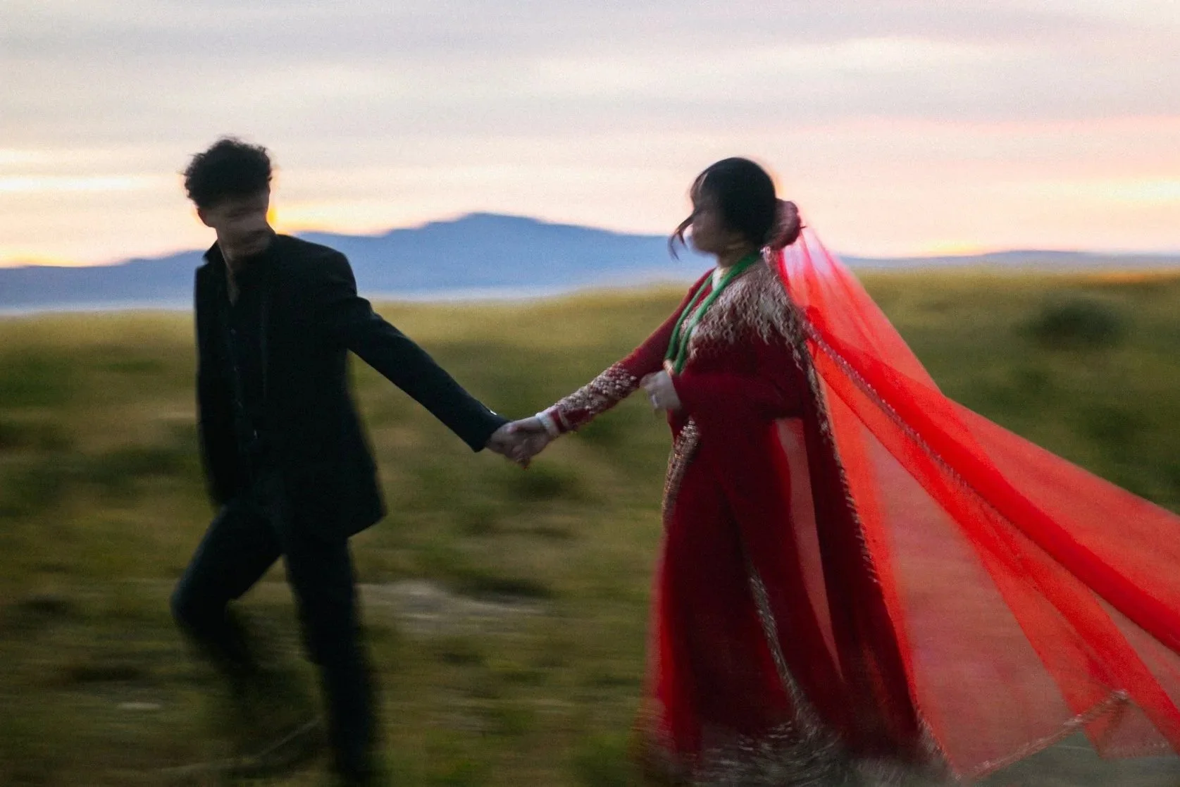 A couple holding hands and walking in a grassy field during sunset, the woman wearing a traditional red dress with a long flowing veil.