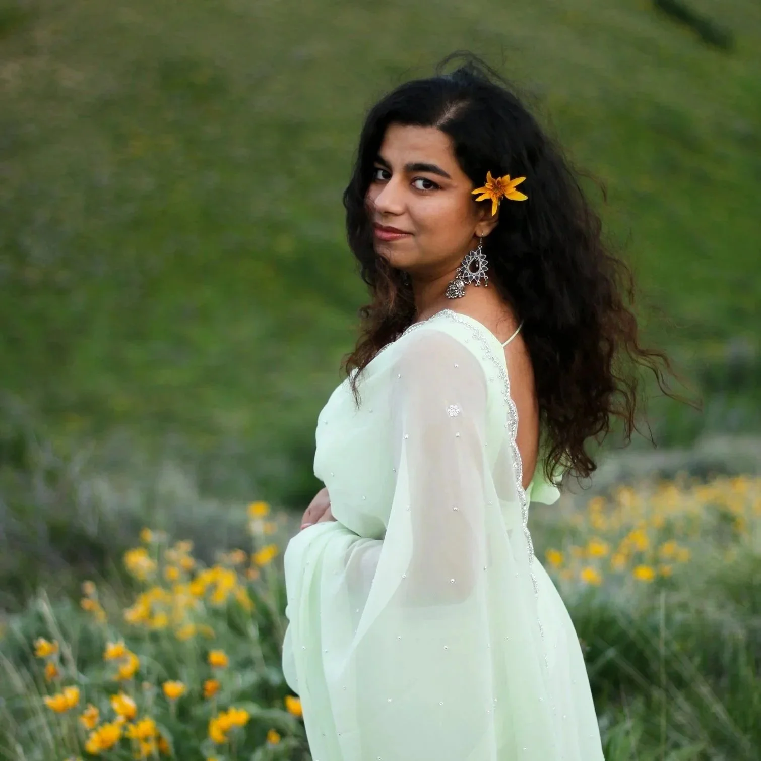 A woman with long, dark, curly hair wearing a light yellow dress with embellishments, standing outdoors in a field of yellow flowers, with a yellow flower in her hair and large dangling earrings.