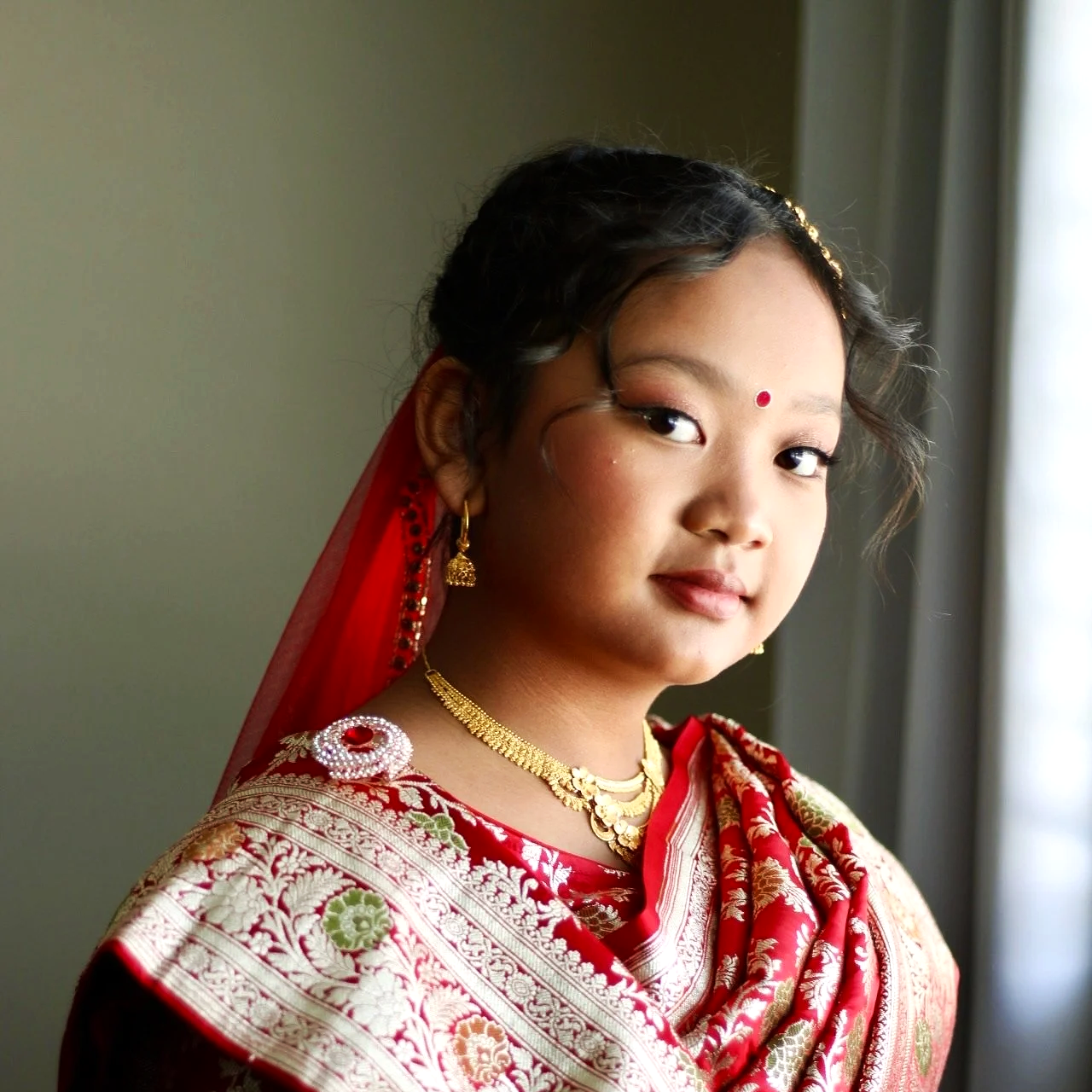 A young girl dressed in traditional Indian clothing, adorned with jewelry, and looking out a window.