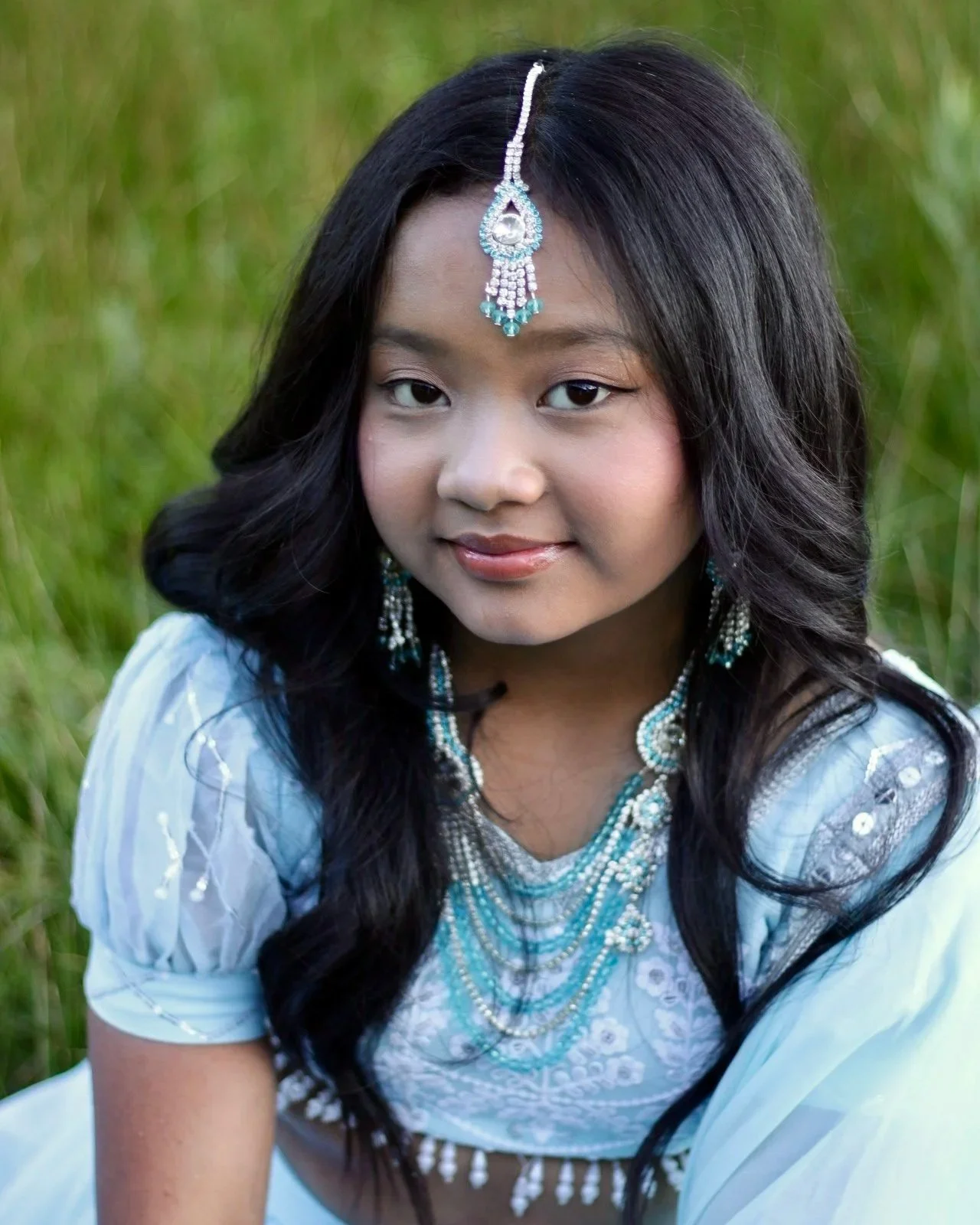 Young girl with long black hair wearing traditional white dress and ornate jewelry, sitting outdoors among green grass.