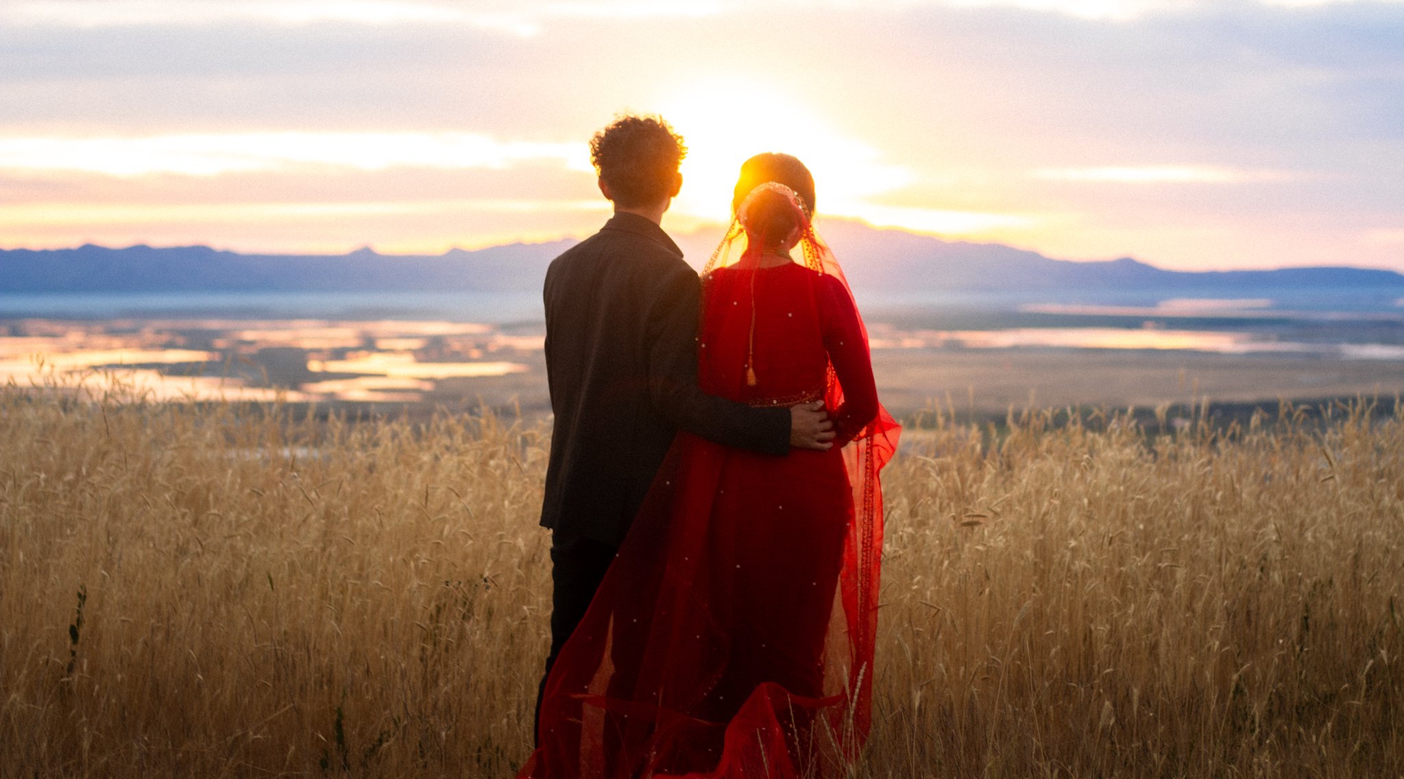 A couple dressed in formal attire, with a woman in a red dress and sheer veil, stands together watching a sunset over a mountain landscape and a lake, embracing each other in a field of tall, dry grass.