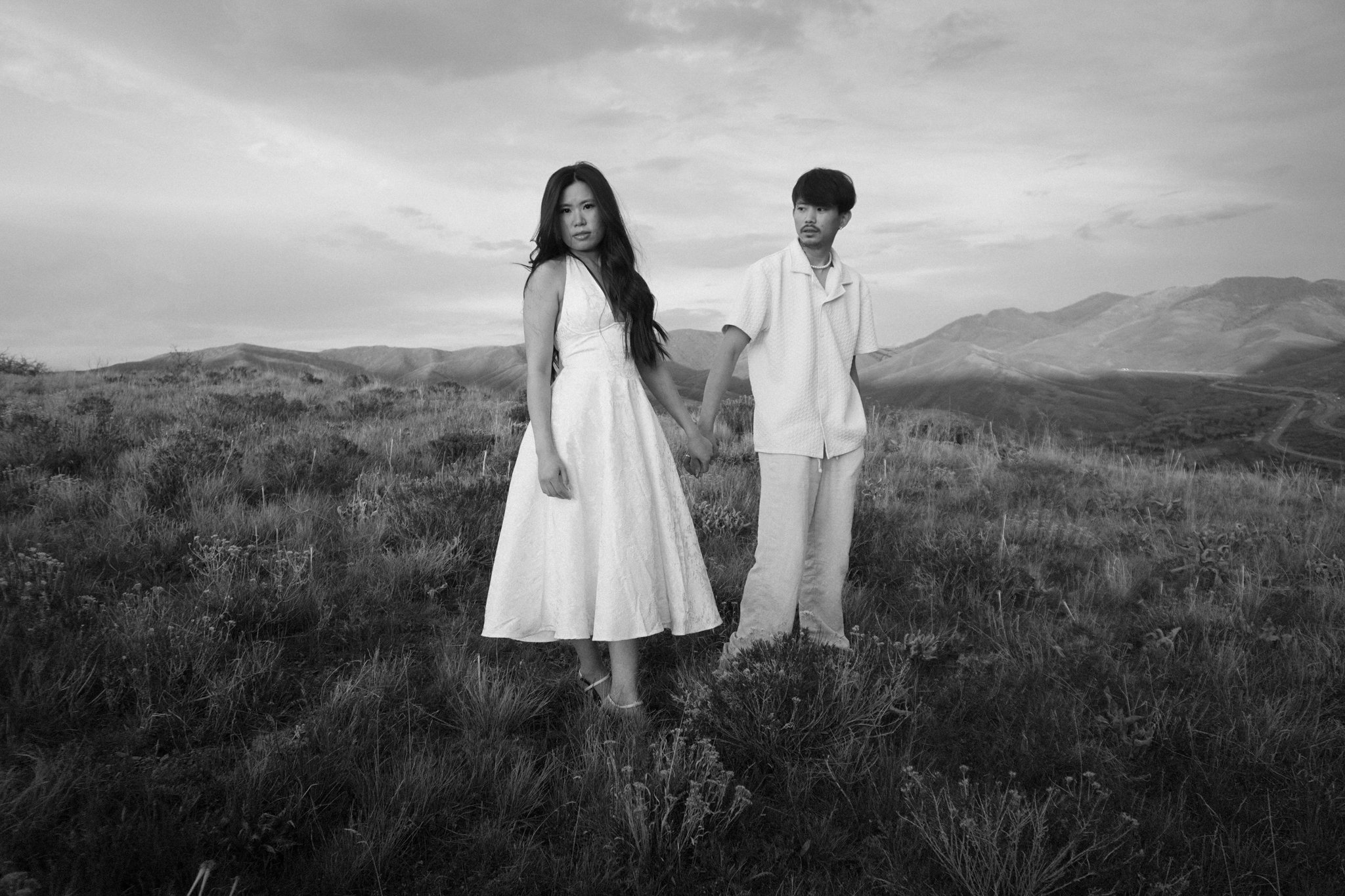 A black and white photograph of a couple holding hands in a grassy outdoor setting, with rolling hills and a cloudy sky in the background.