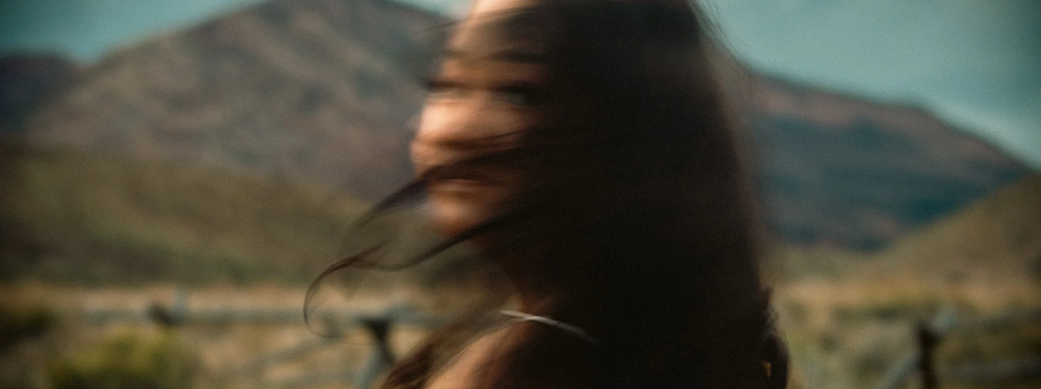 A person with long dark hair standing outdoors in a landscape with mountains and a body of water, wind blowing their hair.