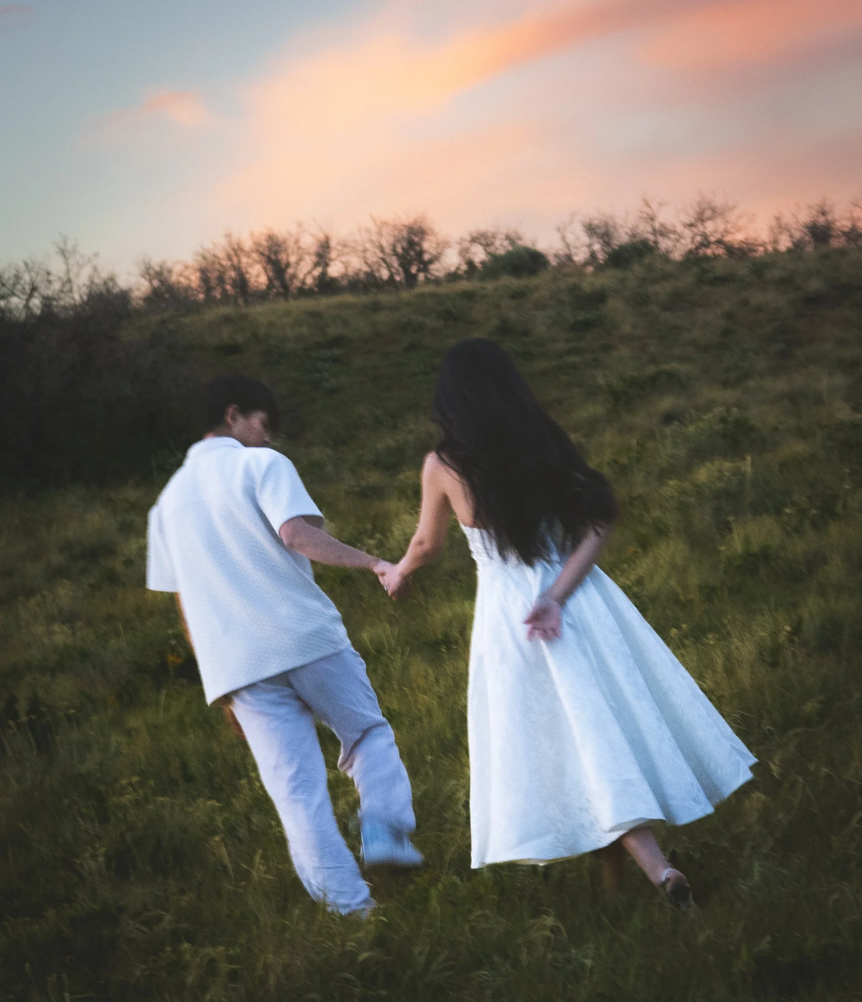 A couple dressed in white holding hands walking on a grassy field at sunset, with a background of trees and a colorful sky.