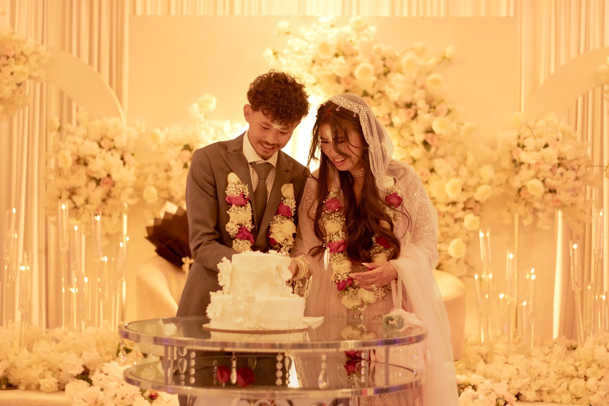 A newlywed couple at their wedding reception, cutting a wedding cake surrounded by floral decorations and soft warm lighting.