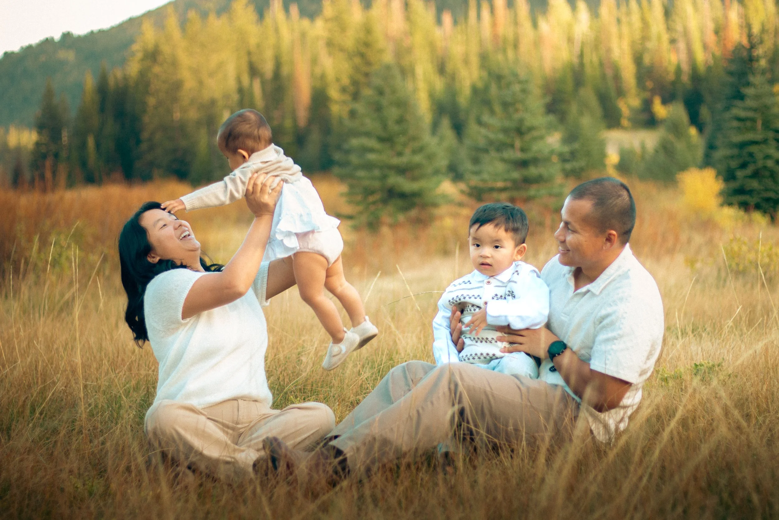 A family outdoors in a grassy field with trees in the background. A woman is holding a child up in the air, smiling. Two young boys and a man are sitting on the grass nearby, watching the woman and child.
