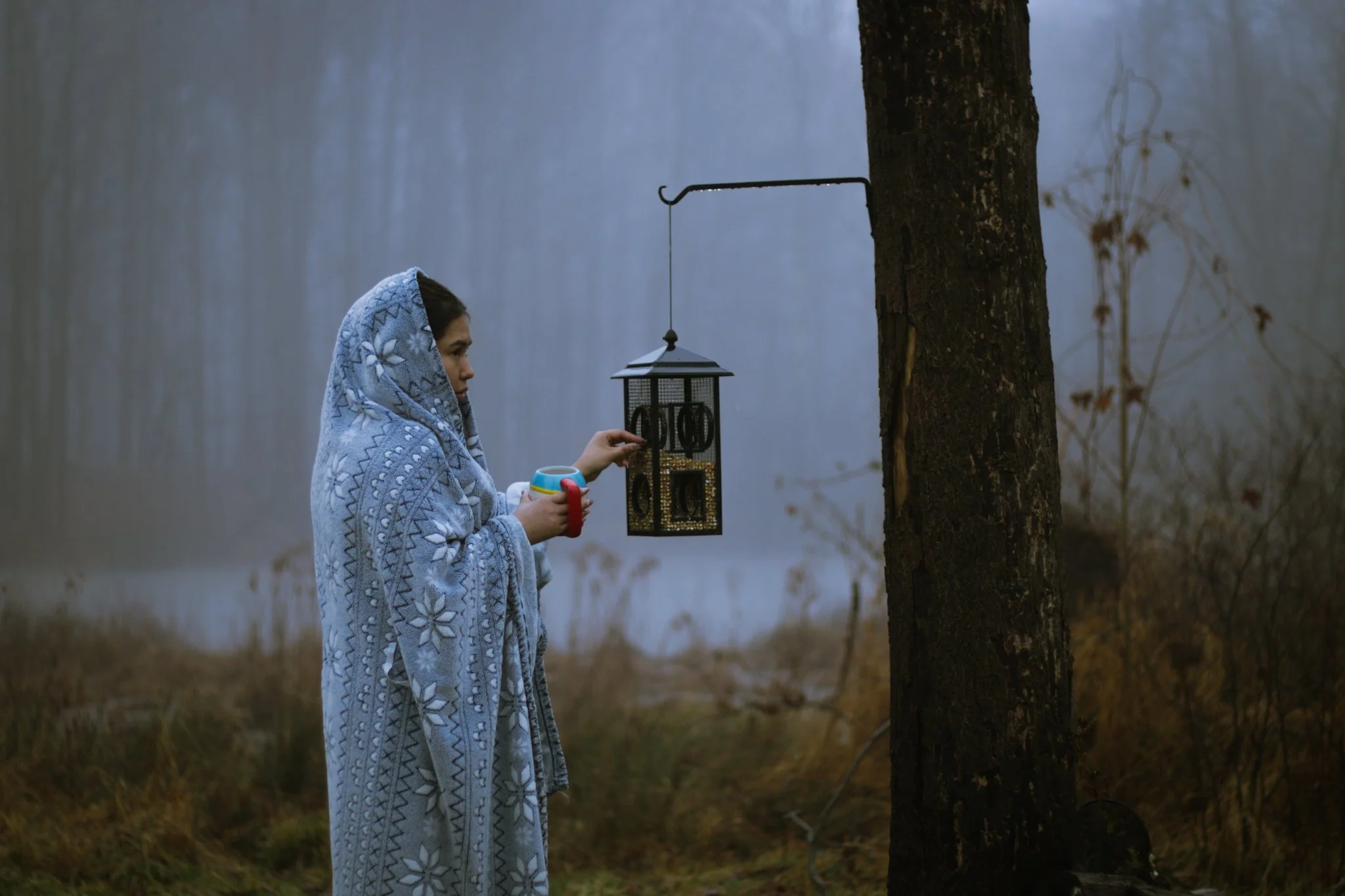A woman in a blue and white patterned shawl is hanging a bird feeder on a tree in a foggy, wooded area. She is holding a mug in her other hand.