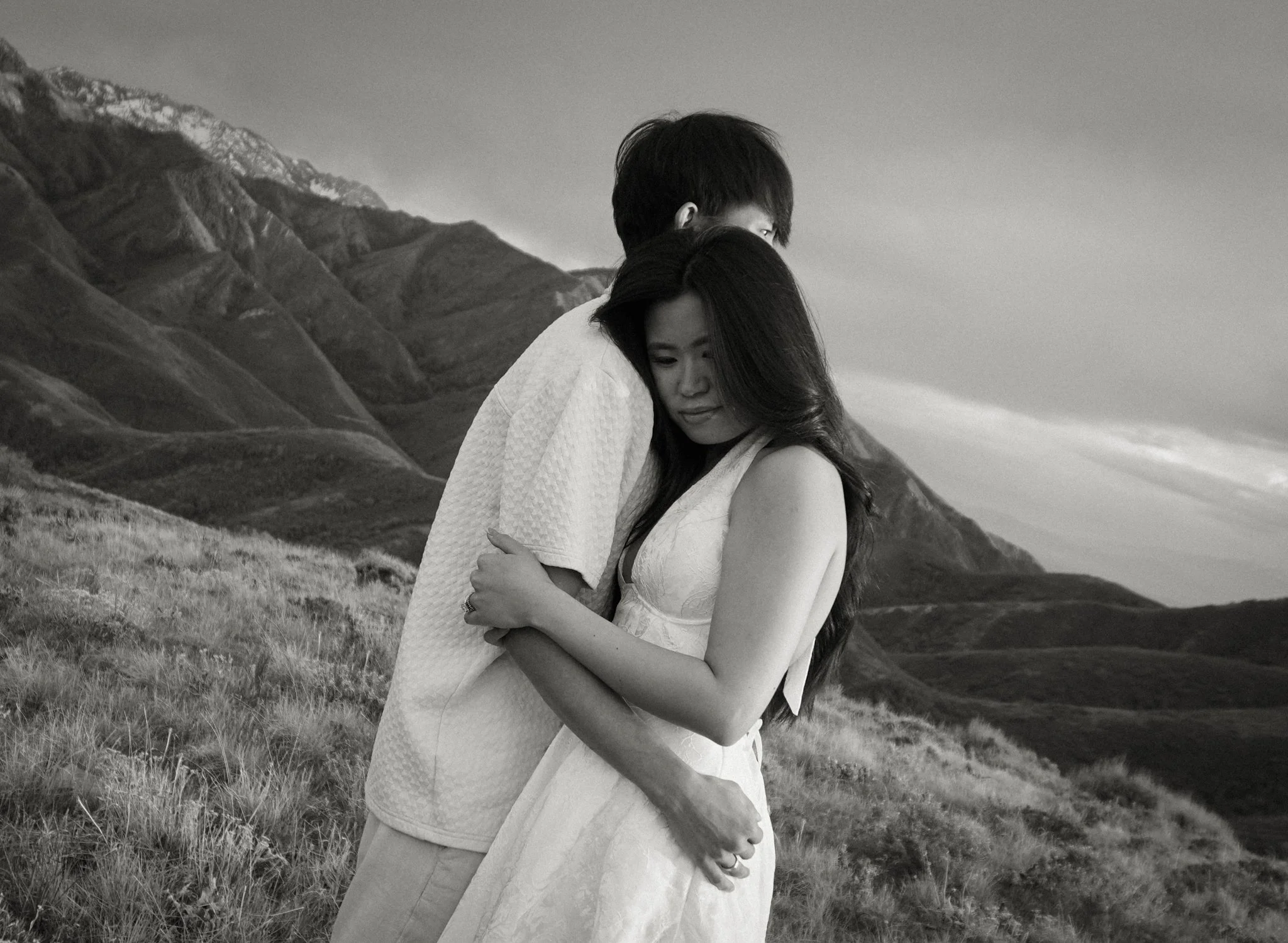 A black and white photo of a young couple standing outdoors on a grassy hill with mountains in the background. They are hugging each other, with the woman in a sleeveless dress and the man in a light-colored shirt. The woman has long dark hair, and t