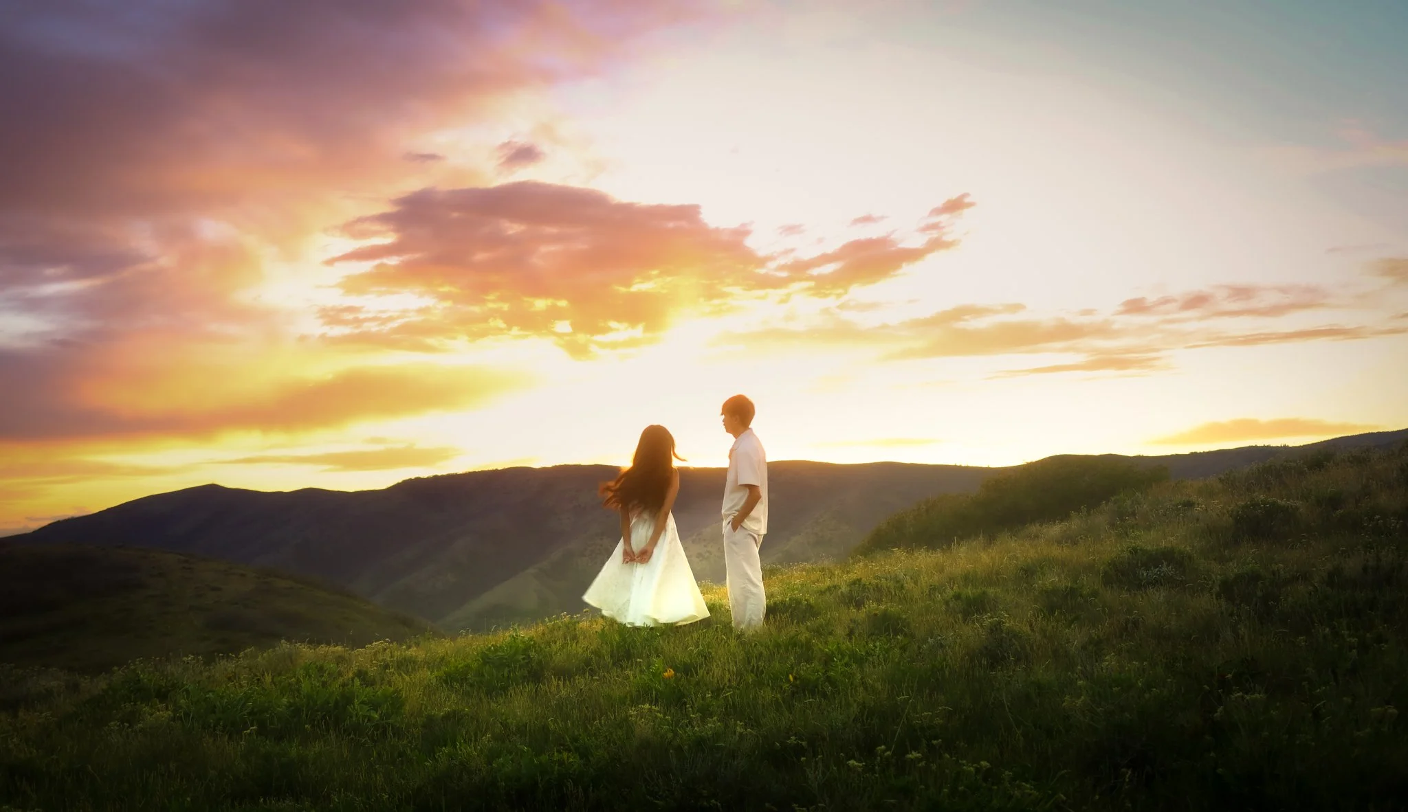A couple stands holding hands in a grassy field at sunset, with a colorful sky and mountains in the background.