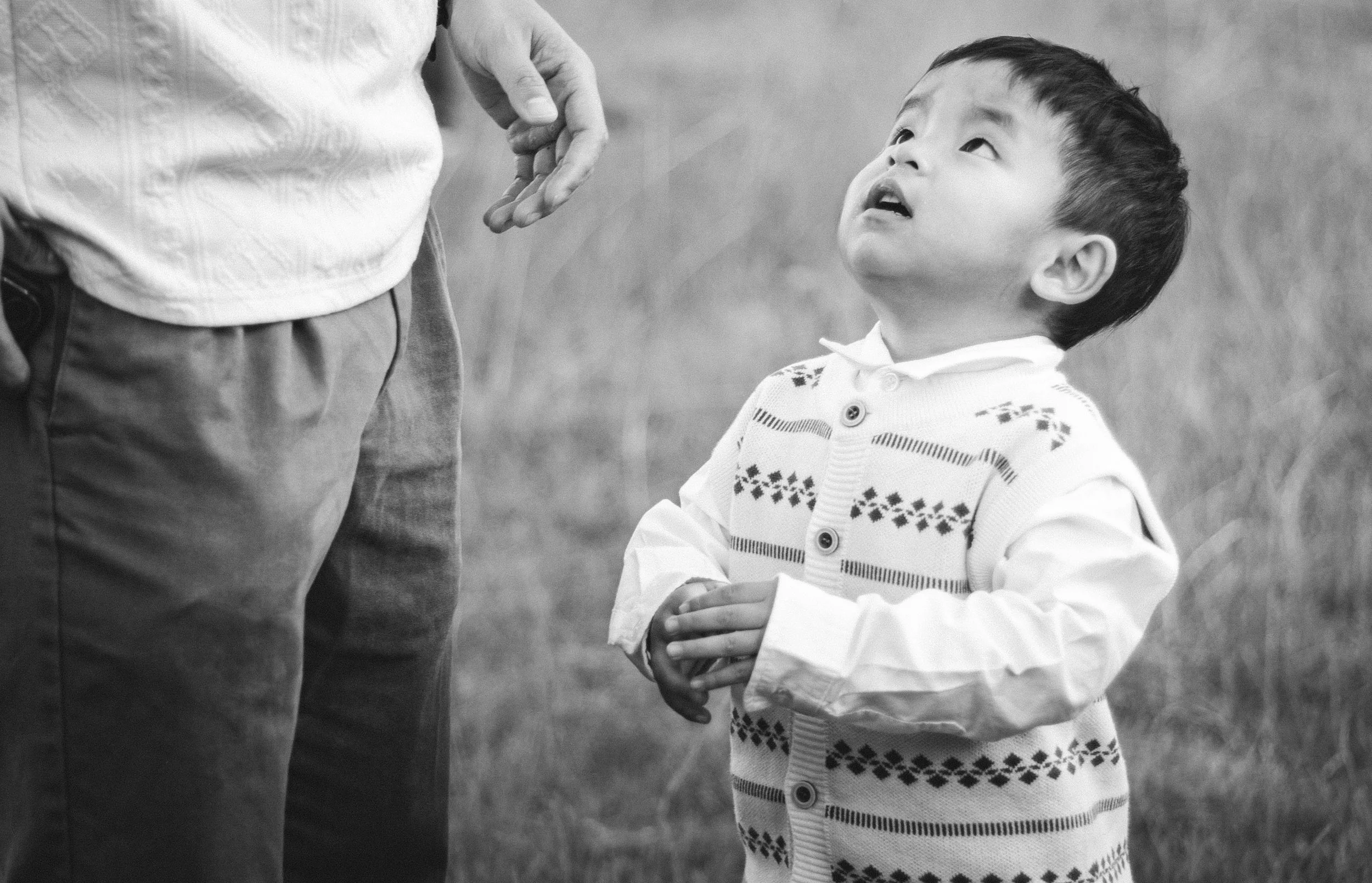A young boy looking up at an adult, holding hands outdoors. The boy is wearing a patterned cardigan. The photo is in black and white.