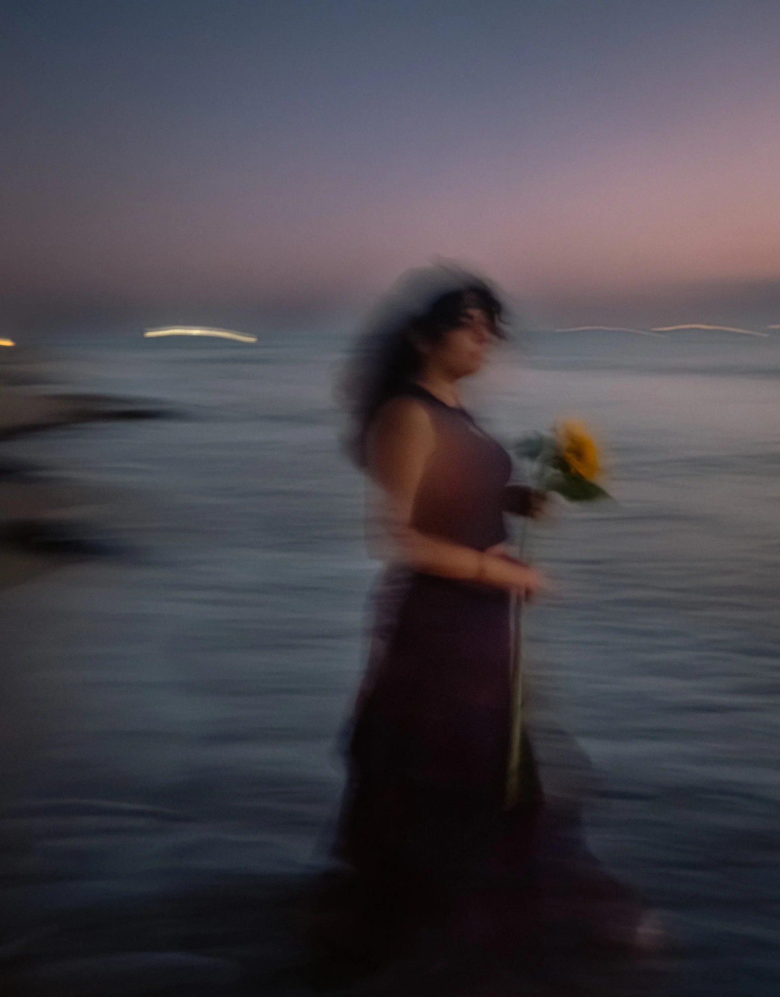 A woman holding a sunflower on a beach during sunset, with the ocean and a colorful sky background, slightly blurred