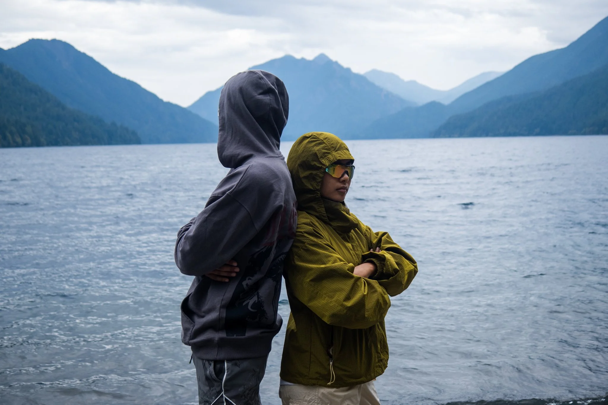 Two people dressed in outdoor gear standing near a body of water, with mountains in the background.