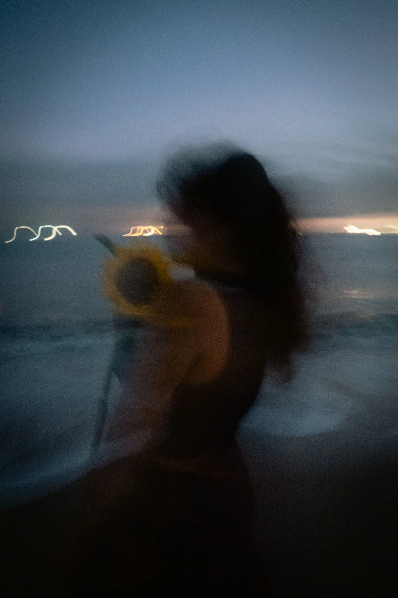 A blurry image of a person holding an umbrella at dusk or night by the water, with distant city lights in the background.