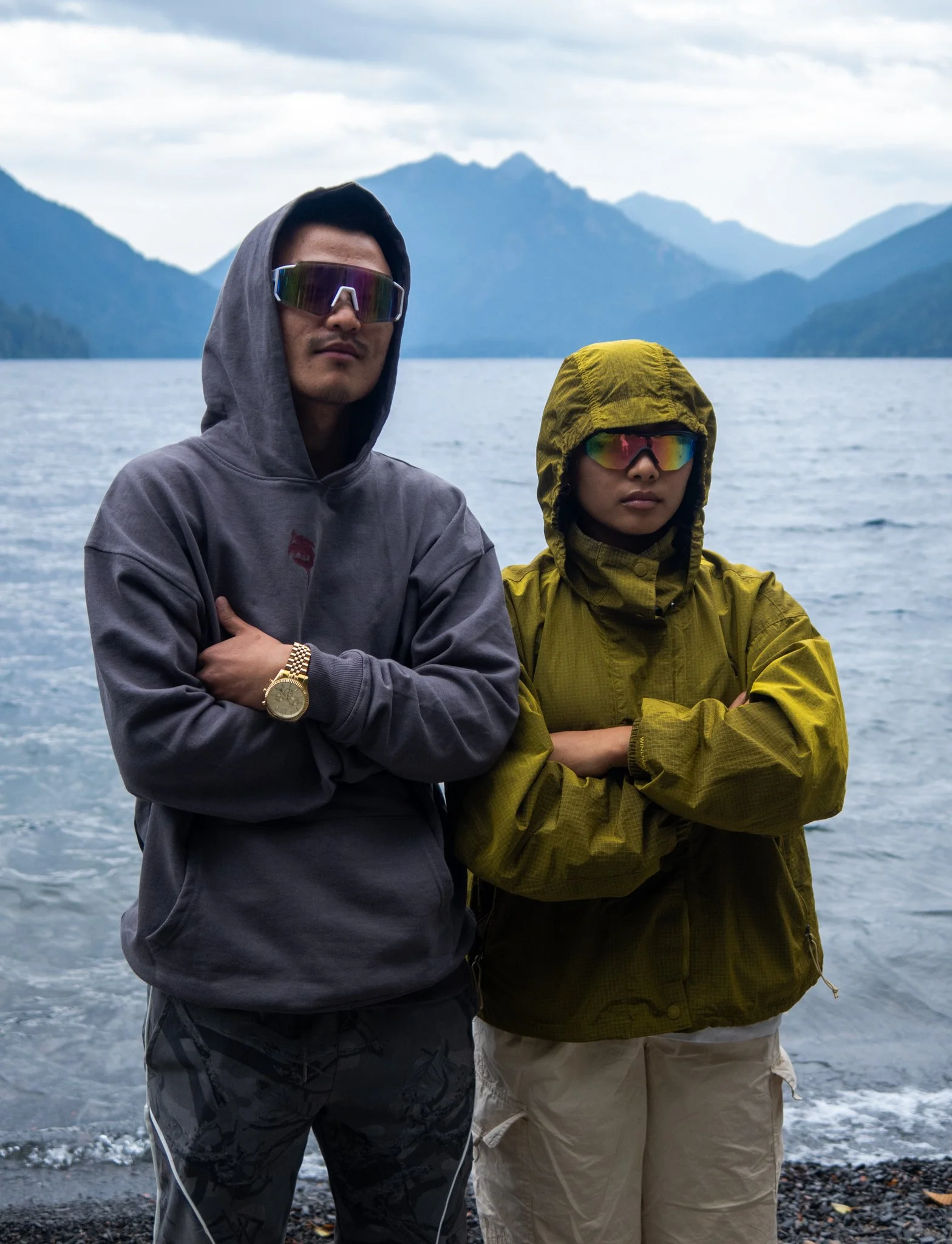 Two men with crossed arms standing in front of a lake with mountains in the background. Both wear sunglasses and hooded jackets, one in gray and the other in yellow-green.