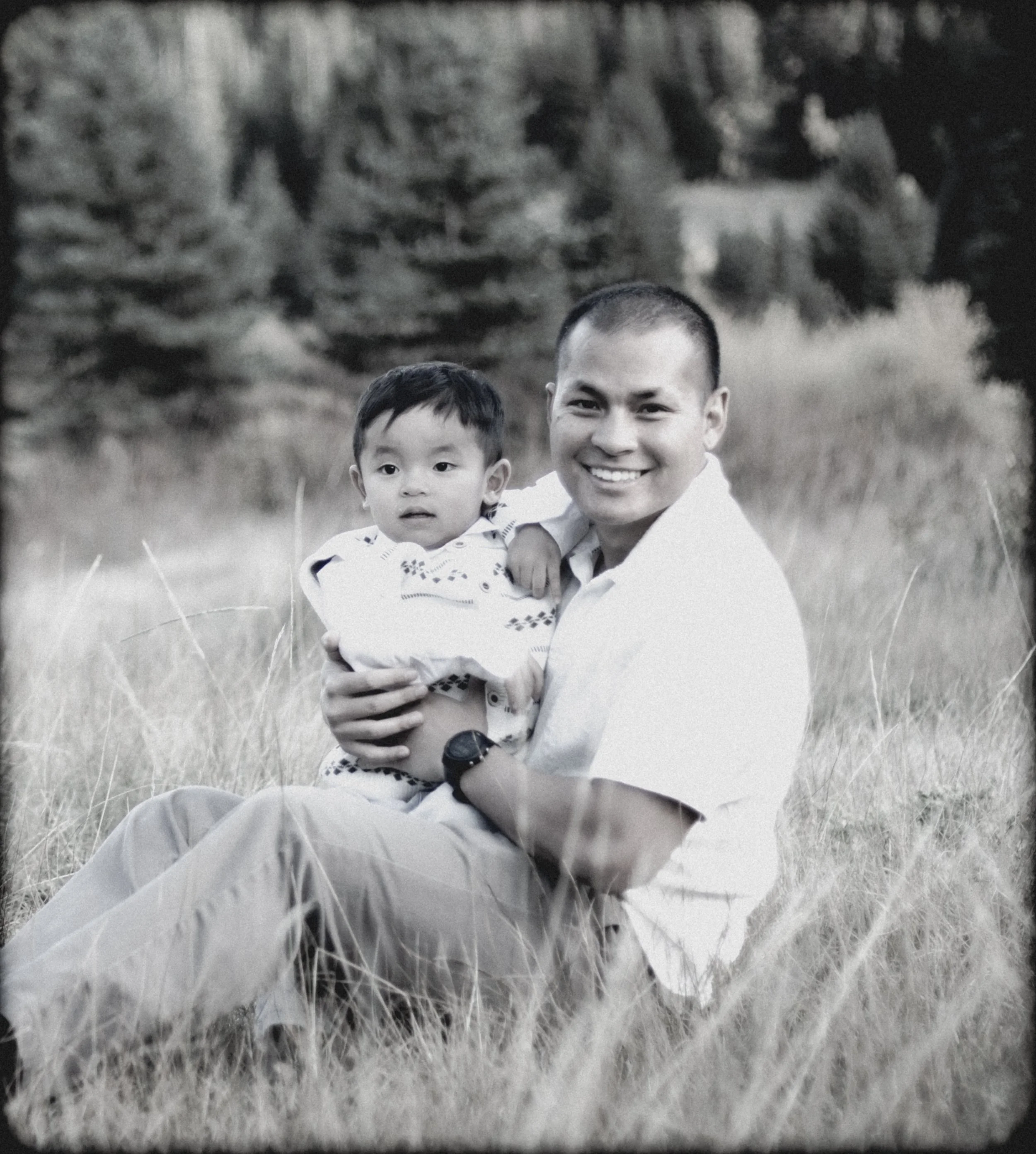 A smiling man holding a young boy outdoors in a grassy field with trees in the background.