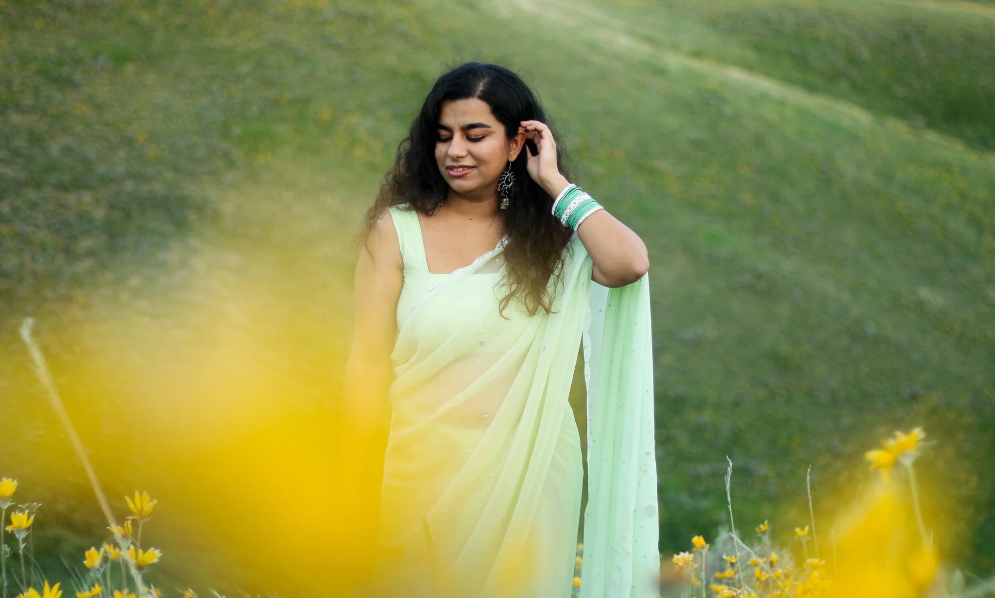 A woman in a light yellow saree standing in a field of small yellow flowers, with her eyes closed and one hand touching her hair, against a green, slightly blurred background.