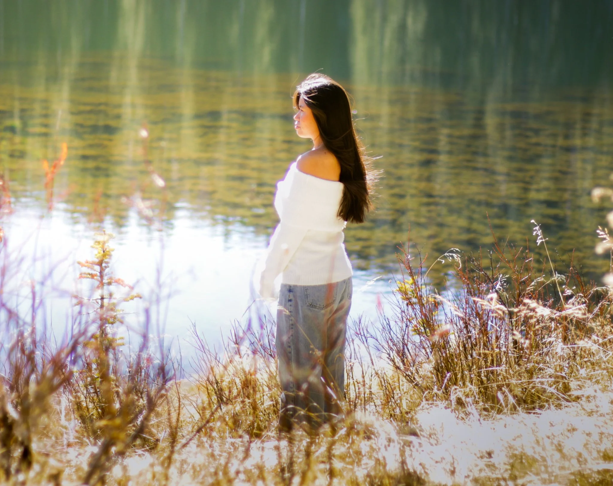 Woman in a white off-shoulder top and jeans standing by a calm lake, surrounded by autumn-colored plants and trees with reflections on the water.