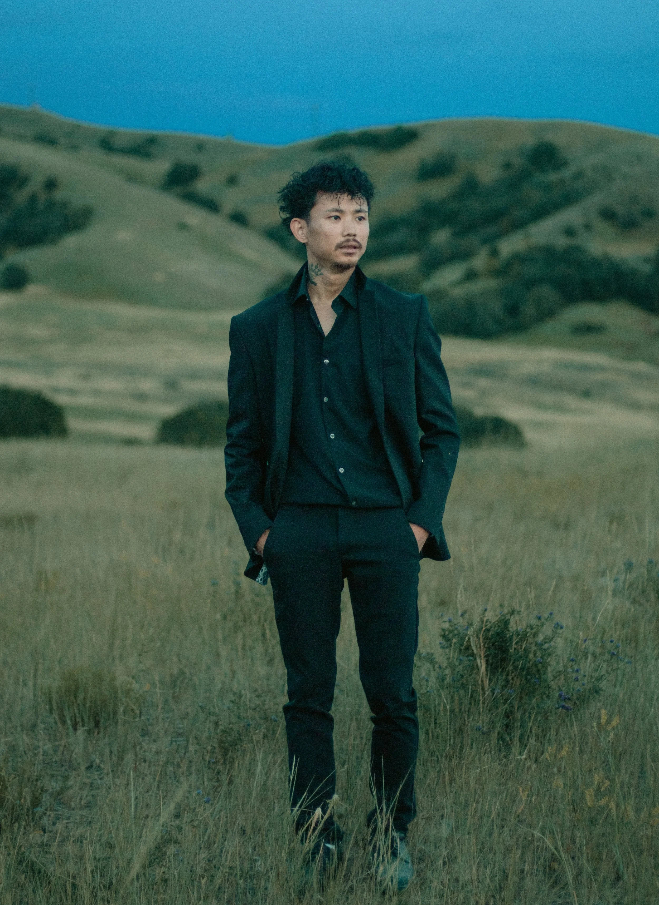 A man in a black suit standing in a grassy field with rolling hills in the background during dusk or dawn.