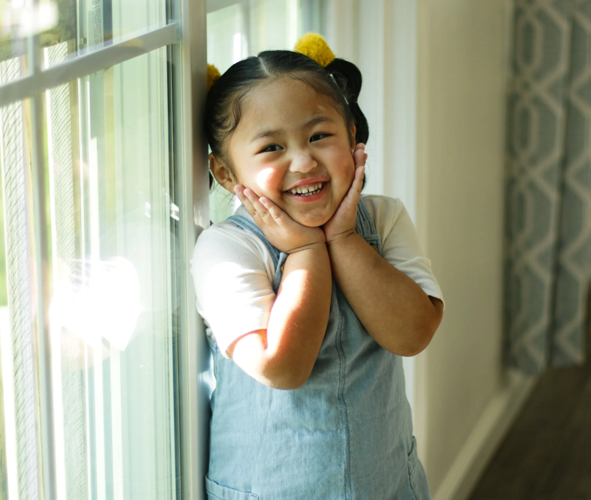 A young girl with pigtails and yellow hair accessories leaning against a window and smiling, with her hands on her cheeks.