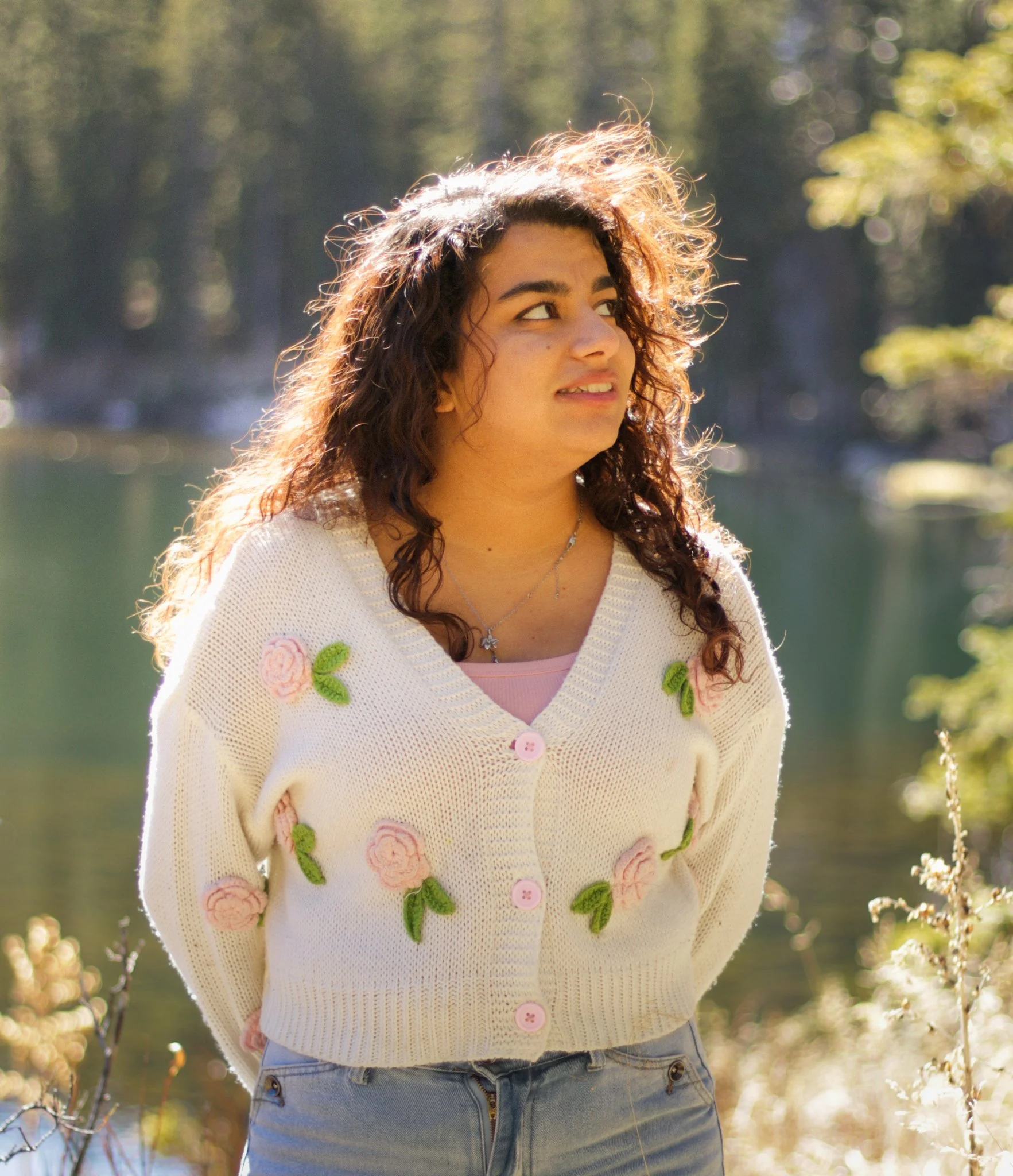 Young woman in a cream sweater with pink and green floral embroidery, standing outdoors in sunlight, with blurred trees in the background.