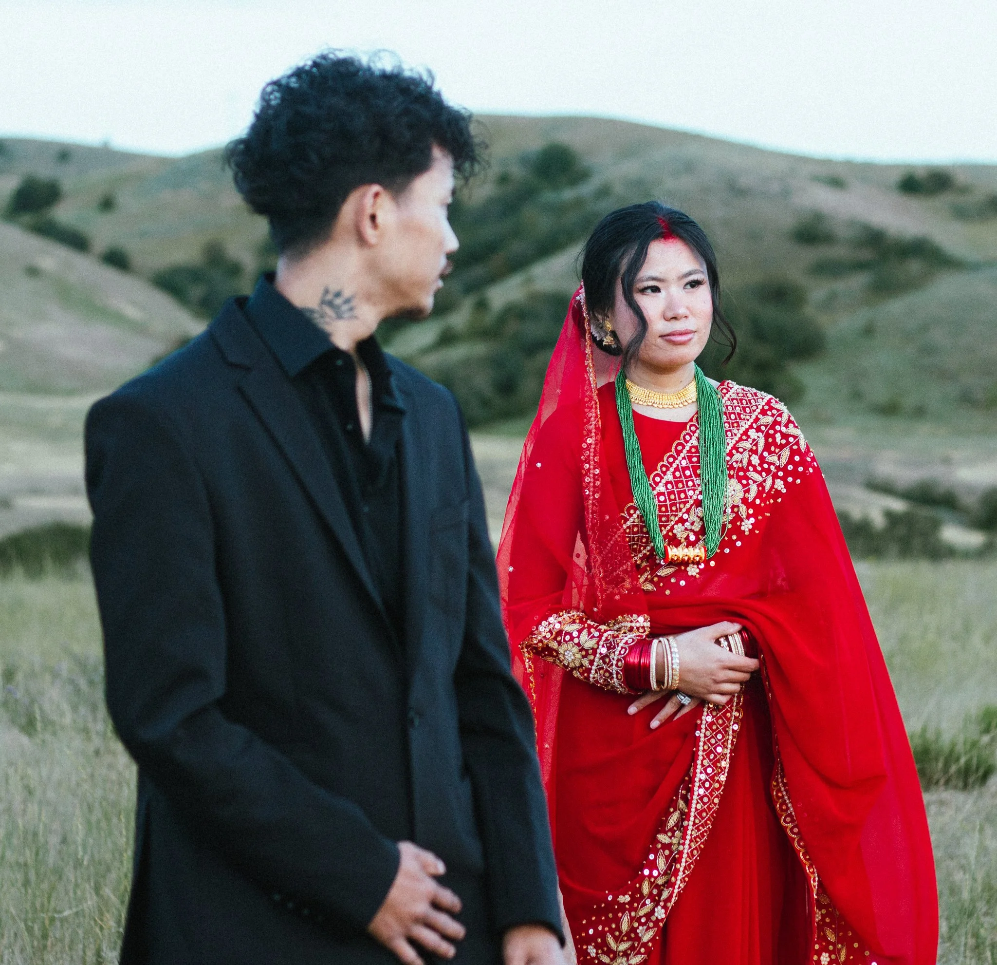 A man and woman dressed in traditional South Asian attire standing outdoors in a grassy, hilly landscape.