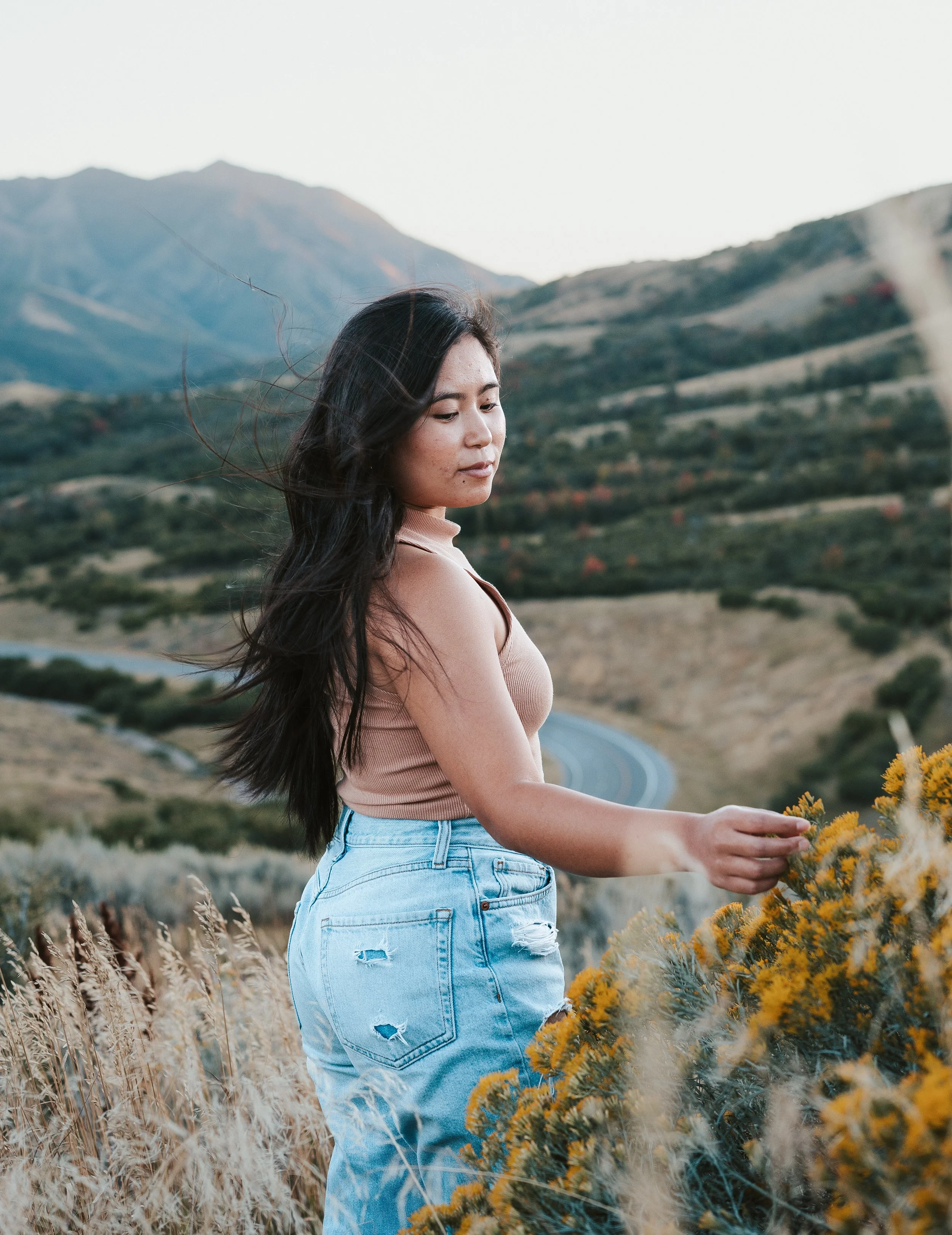 A woman with long dark hair wearing a beige top and ripped light blue jeans standing in a field of yellow flowers with mountains in the background