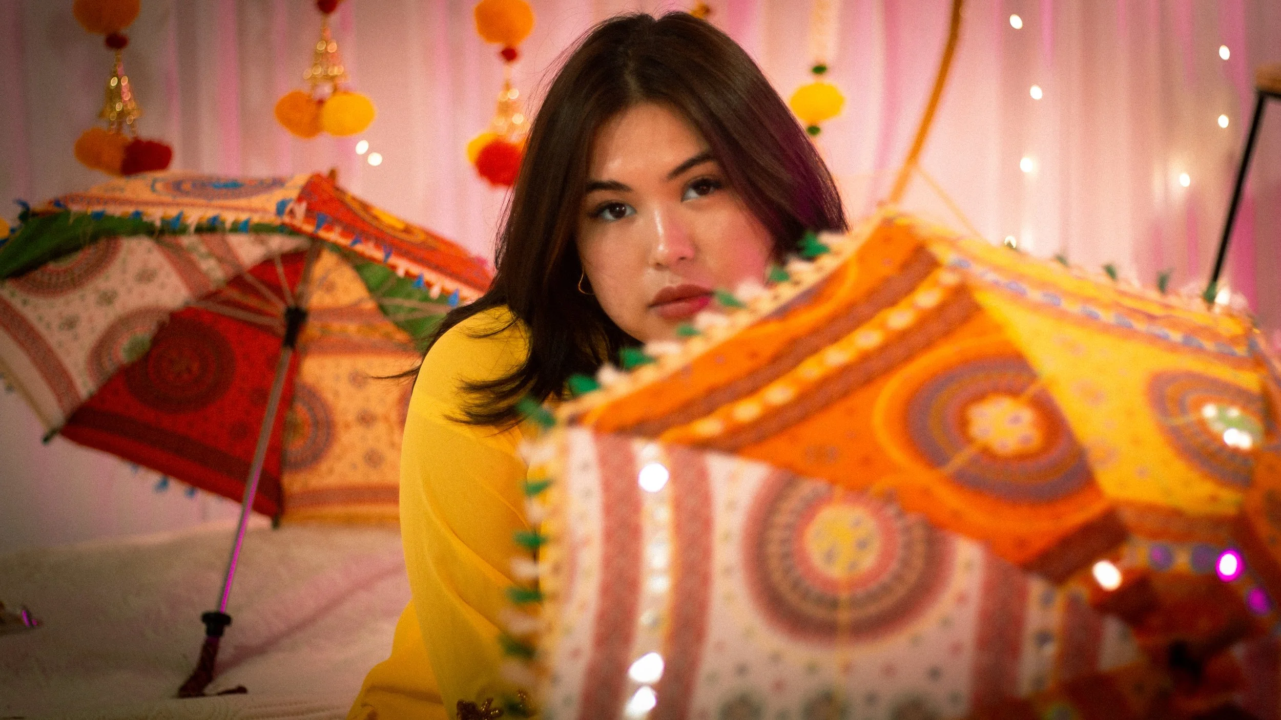 A woman with dark hair wearing a yellow top, surrounded by colorful patterned umbrellas and hanging lanterns, in a vibrant, decorative indoor setting.