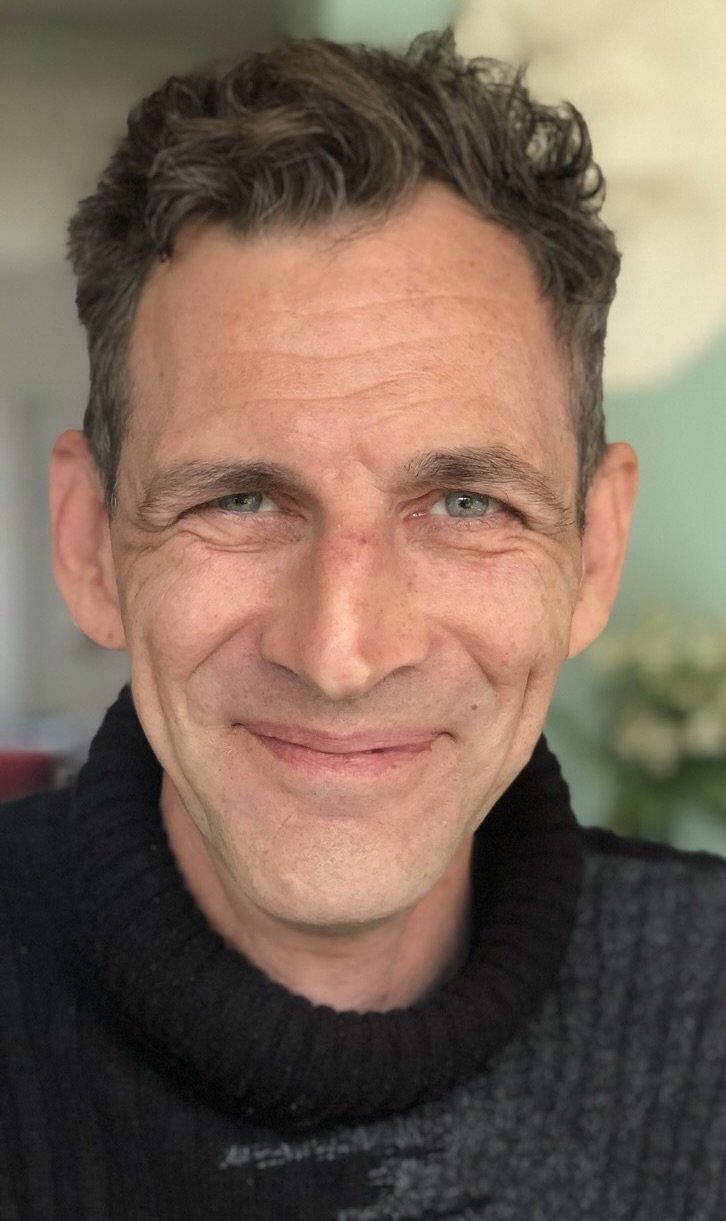 Close-up of a smiling middle-aged man with short, curly dark hair, blue eyes, and light skin, wearing a black turtleneck sweater.