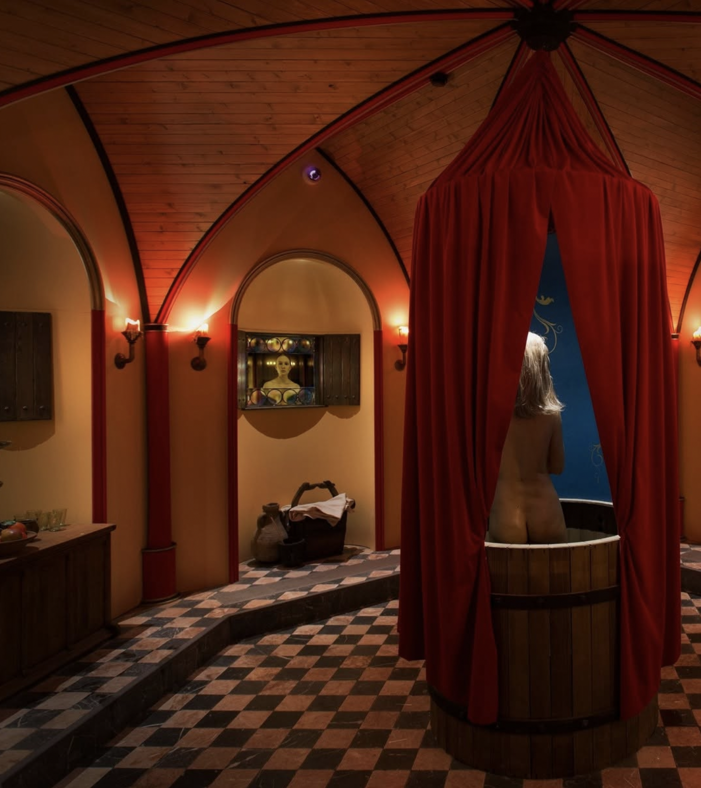 A woman with blonde hair is standing in a circular wooden tub, partially covered by red curtains in a warmly lit room with wooden ceiling and patterned tile floor. The room has a vintage, cozy feel.