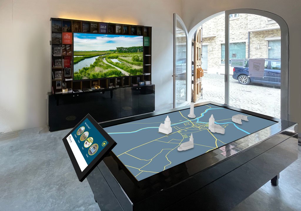 An indoor exhibit with a 3D physical map of a city or area, featuring white model buildings, on a raised platform with a digital informational display. In the background, a large TV screen shows a landscape with water, greenery, and a path, with a bo