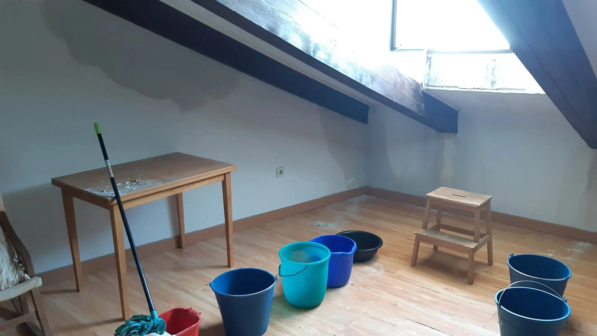 Habitación en proceso de limpieza o mantenimiento con cubetas de agua de varios colores, un trapeador y una mesa pequeña. La habitación tiene un techo inclinado con una ventana grande que deja entrar mucha luz.