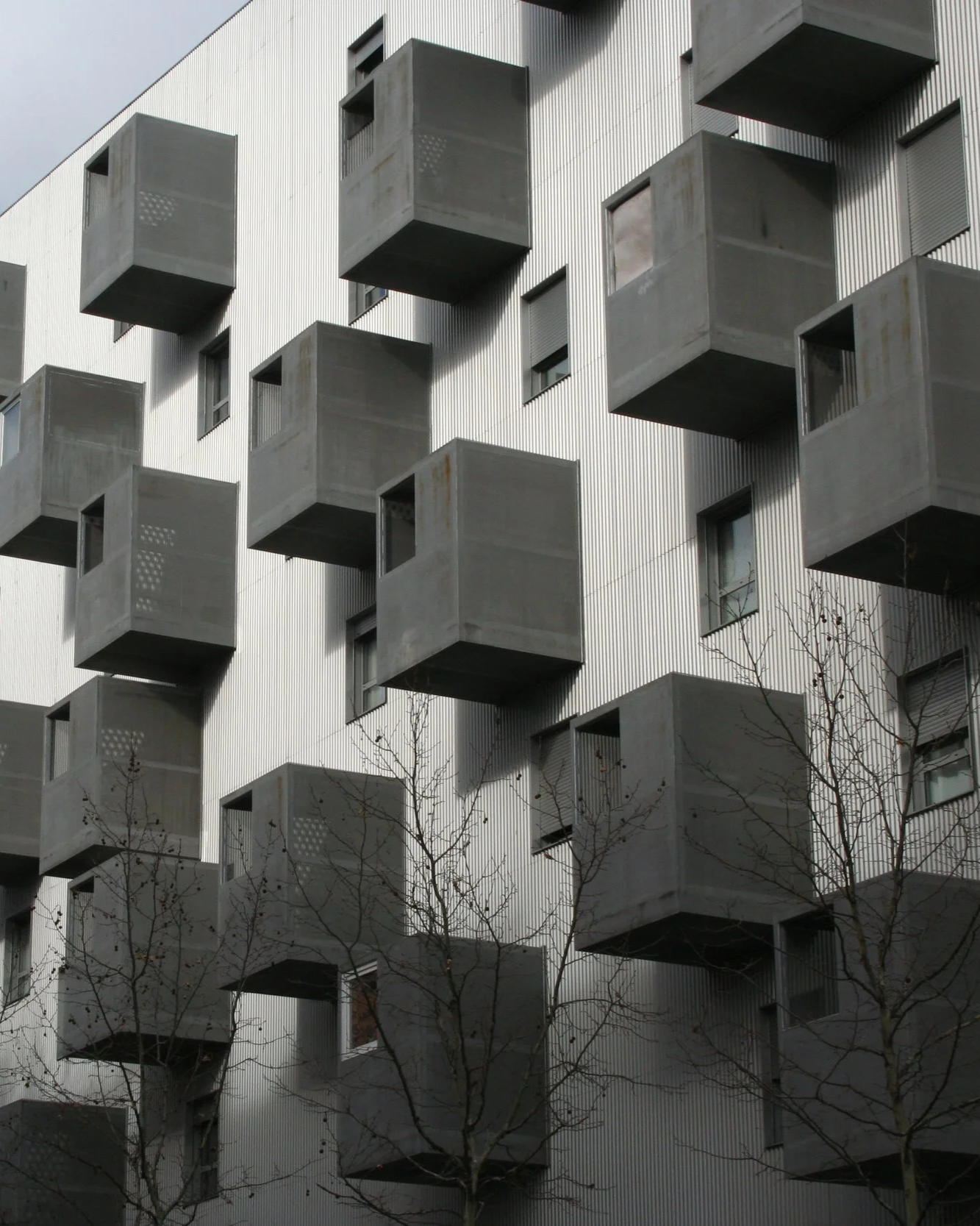 Edificio moderno con ventanas en forma de cajas de concreto que sobresalen y crean patrones geométricos. Hay árboles sin hojas en el frente.