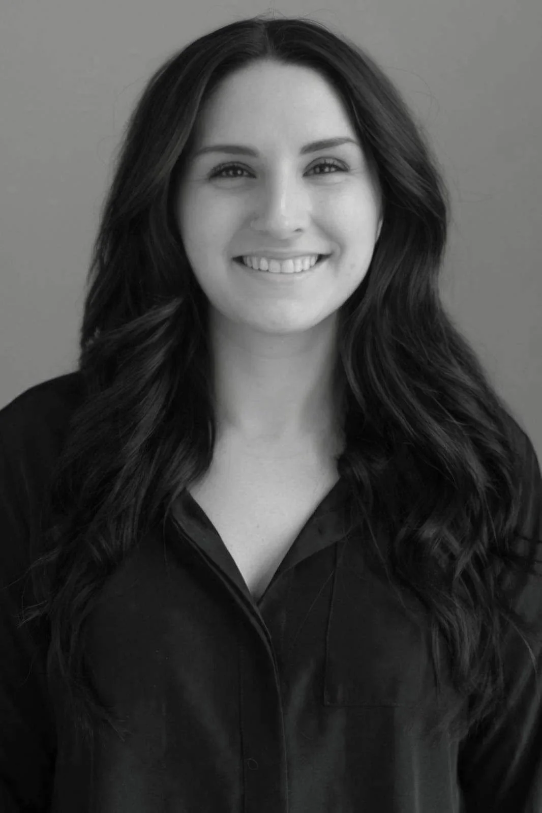 Black and white portrait of a woman with long, wavy hair smiling at the camera, wearing a dark collared shirt.