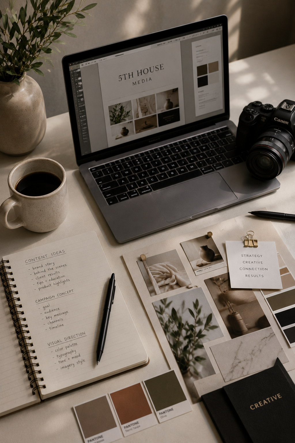 A workspace with a laptop displaying a design project for 5th House Media, a camera, a notebook with content ideas, photographs, color samples, a cup of coffee, and decorative items.