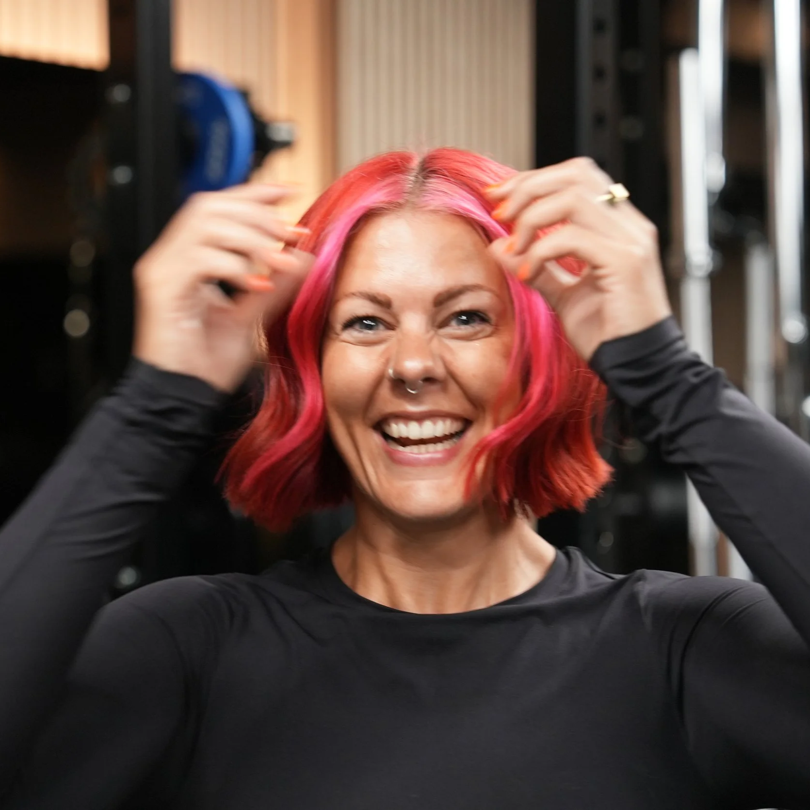 A woman with pink and orange hair, smiling happily, adjusting her hair in a salon or studio setting.